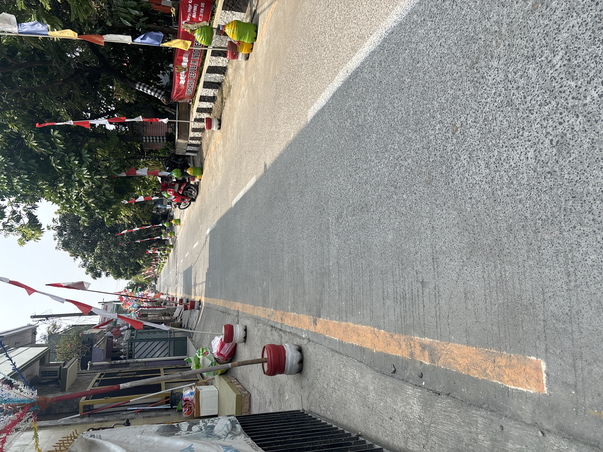 The image depicts a street scene in Telagamurni, Indonesia, decorated with Indonesian flags and pennants. The flags are red and white, indicating national celebrations. The street is paved with asphalt and has a yellow line marking the edge of the sidewalk. On the left side of the street, there is a sidewalk with some buildings and a fence. Various decorative items, including colorful pennants on sticks, are placed along the sidewalk. Some of these pennants are made from recycled plastic bottles painted red and white. On the right side of the street, trees line the road, and more flags and decorations are visible. A red banner with text in Indonesian, likely related to a national holiday, is also present. Several motorcycles are parked along the right side of the street, and large, colorful plastic containers, also decorated, are positioned at intervals along the edge of the road. The overall impression is one of festive decoration and community engagement.