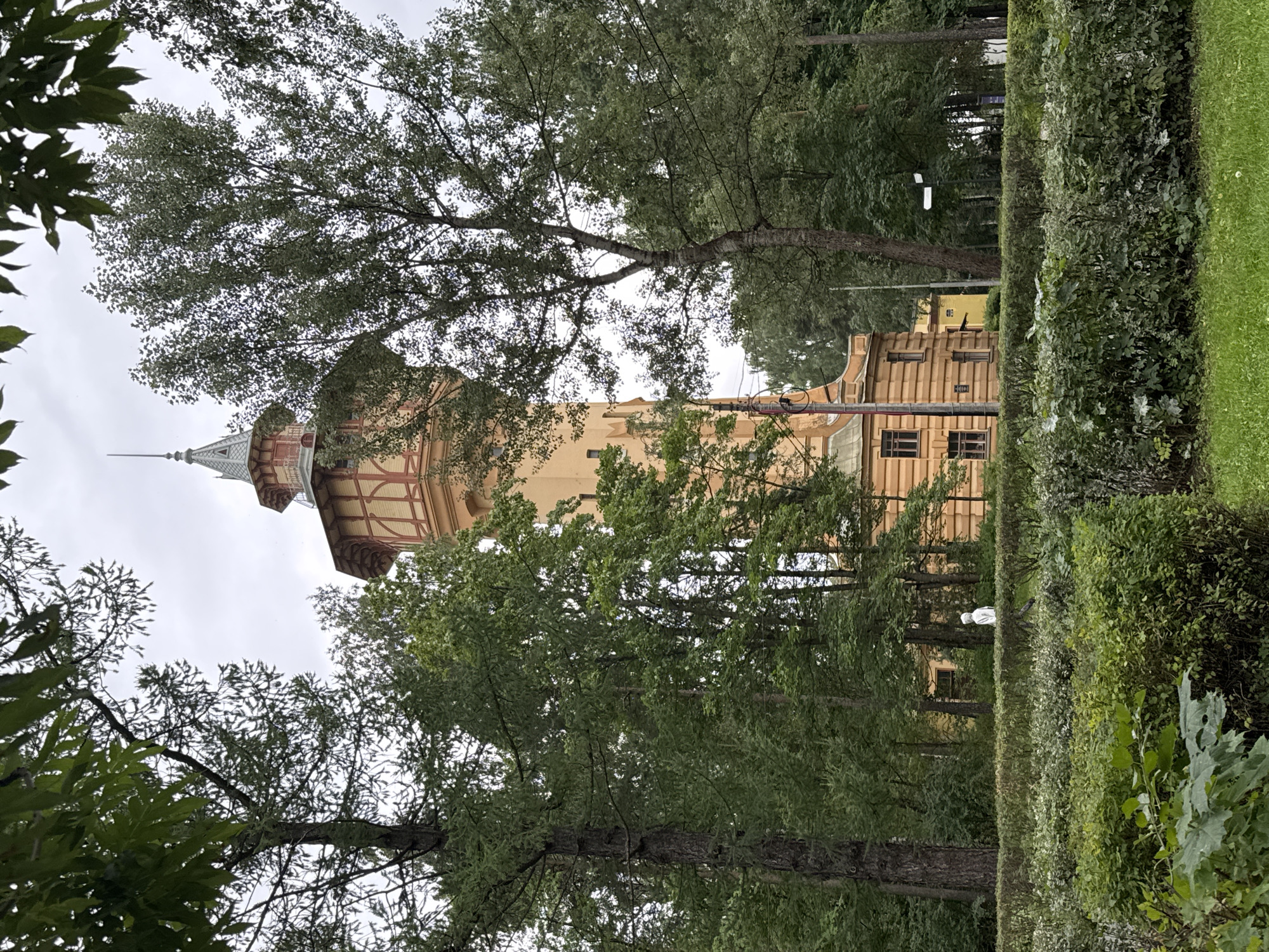A tall, yellow tower with red trim stands among lush green trees, partially obscured by their foliage. The tower has a pointed roof and windows on its upper levels. In the foreground, a neatly trimmed hedge separates a grassy area from the rest of the scene. A single person wearing a white hooded jacket walks in the middle ground, facing away from the camera. The sky is overcast. This scene is in the city of Ladozhskoe Ozero, Russia.
