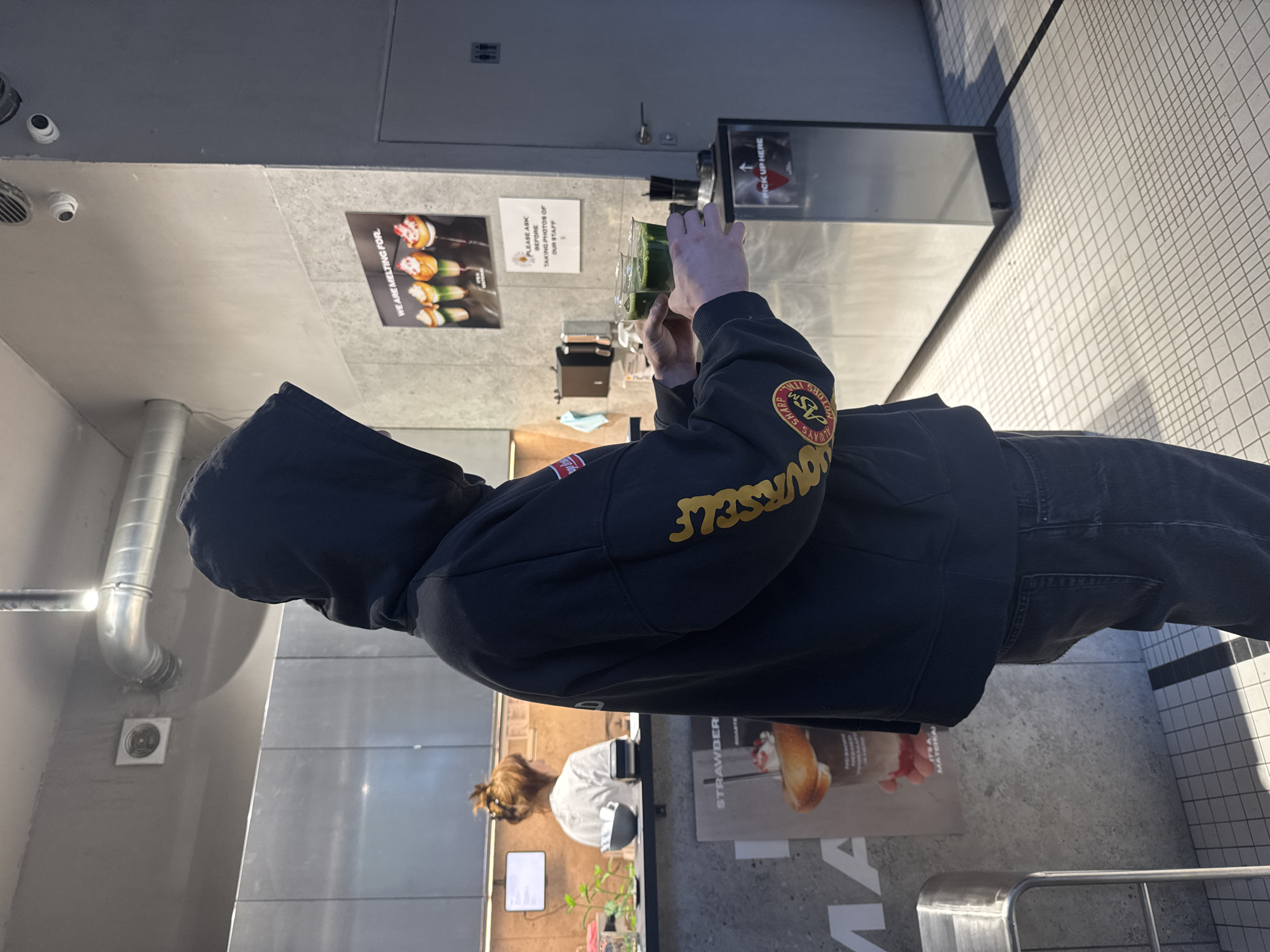 A person, viewed from behind and slightly to the side, stands in what appears to be a modern cafe or dessert shop in Warsaw, Poland. The individual is wearing a dark blue or black hooded sweatshirt with yellow text on the right sleeve that reads, "ALWAYS SHARP ASM MOTORS YOURSELF" (partially inverted). They are also wearing dark jeans. Their hands are holding a transparent glass containing a vibrant green beverage, possibly matcha, and they are simultaneously holding a smartphone, suggesting they are photographing or videoing the drink.
The interior of the establishment features light grey, concrete-like walls and a white tiled floor with a grid pattern, illuminated by bright overhead lighting and possibly some natural light casting shadows. In the background, behind a counter, a staff member with hair tied up is partially visible.
Several signs and posters are displayed on the walls. One poster advertises various desserts or drinks with the text "WE ARE MELTING FOR". Another sign clearly states, "PLEASE ASK BEFORE TAKING PHOTOS OF OUR STAFF", indicating a policy within the venue. A "PICK UP HERE" sign with an upward arrow is also visible, marking a service point. Metallic ventilation ducts are attached to the walls and ceiling, alongside security cameras. On the counter, a small potted plant adds a touch of greenery. In the foreground, part of a metal railing is visible. The scene captures a typical moment in a contemporary urban eatery, with a customer documenting their purchase.