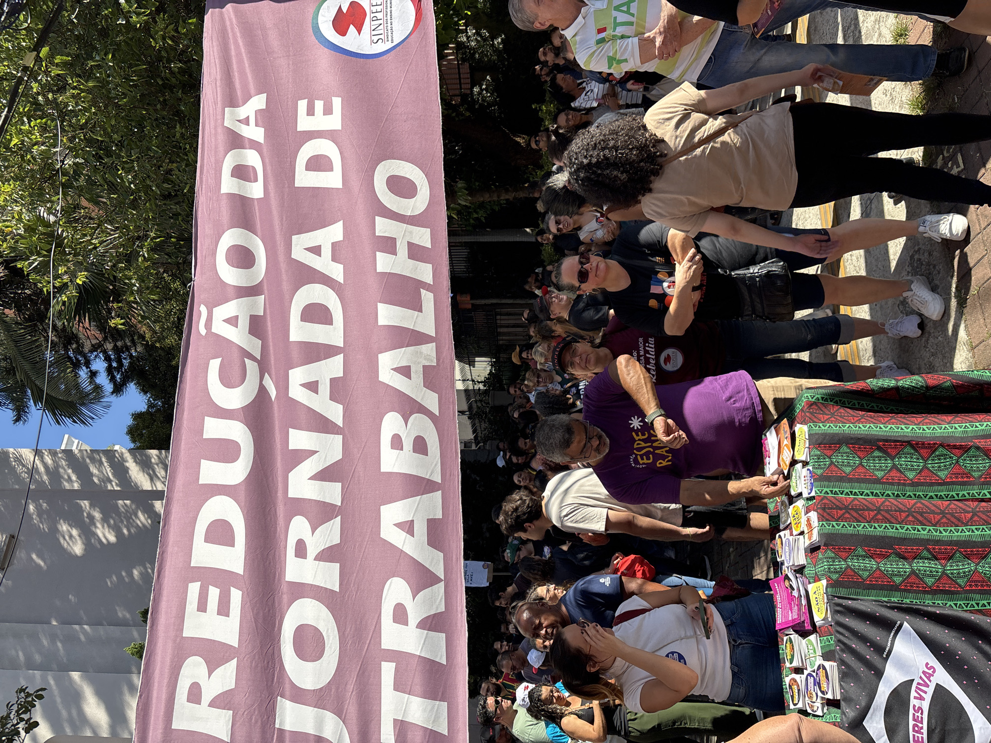 The image shows a daytime outdoor scene in São Paulo, Brazil, featuring a demonstration advocating for the reduction of working hours. A crowd of people is gathered around a table displaying pamphlets and stickers, suggesting an organized event or rally. Prominently displayed is a large, lavender-colored banner with white lettering that reads "REDUÇÃO DA JORNADA DE TRABALHO," meaning "Reduction of the Workday." The logo of "SINPEE" is also visible on the banner. A portion of another flag with the words "ERES VIVAS" can be seen. People of various ages and ethnicities are visible, engaged in browsing the materials on the table, interacting with one another, and participating in the demonstration. The environment includes trees and buildings in the background, indicating an urban setting.