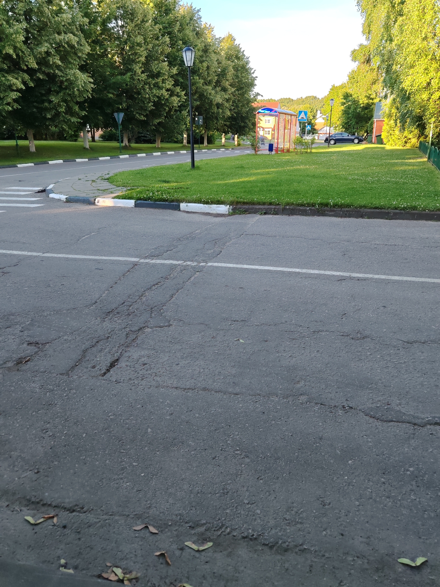 This media file depicts a street scene in Rosinka, Russia, likely during the daytime. The foreground is dominated by a cracked asphalt road with a white line marking a lane. A pedestrian crossing is visible on the left side of the road, marked with white stripes. The road curves to the left, bordered by a curb with black and white markings, and a well-maintained grassy area with scattered white flowers.
Beyond the grass, a row of mature trees with lush green foliage lines the street. A classic black lamppost stands on the grassy median. Further into the scene, a small bus stop shelter with a red and yellow frame and a translucent roof is visible. To the right of the shelter, a dark blue sedan is parked on the grass. In the background, there are buildings and more trees, with a slightly hazy sky suggesting it might be late afternoon or early morning. The overall impression is one of a peaceful, suburban or semi-rural environment on a clear day. There is no discernible text in the image.