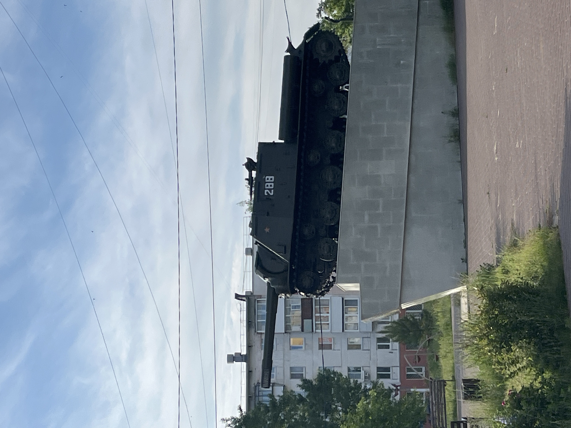 The image displays a dark green military tank mounted as a monument on a large, textured concrete block pedestal in an urban setting. The tank, likely a Soviet-era self-propelled artillery piece or tank, features the white number "288" prominently on its turret and a red star emblem. Its long gun barrel extends horizontally towards the left.
In the background, multi-story residential buildings, consistent with apartment blocks, are visible with numerous windows. Green trees and bushes are located around the base of the monument and in front of the buildings. To the right, a paved area, possibly a sidewalk or road, is partially visible. Overhead, multiple power lines crisscross against a clear blue sky with scattered clouds.
The scene is captured during bright daylight, suggesting late morning or afternoon with sunny weather. No people or activities are visible, emphasizing the static nature of the monument. The general architecture and the tank's design are consistent with its location in Chaikovsky, Russia. The only visible text is the number "288" on the tank.
