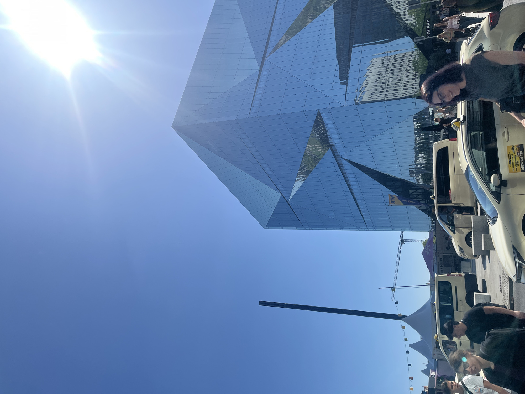 The image depicts a sunny day in Berlin, Germany, outside a modern, angular glass building. The building's facade is highly reflective, mirroring the sky and surrounding structures. In the foreground, several people are visible walking and standing. A yellow taxi with the words "TAXI FAHRER GESUCHT!" and a phone number is prominently featured. Yellow vans, possibly taxis or shuttle vehicles, are also parked in the area. In the background, tents, a crane, and other buildings can be seen, suggesting an urban environment with ongoing construction or an event. The sun is bright and high in the sky, casting lens flares and illuminating the scene.
