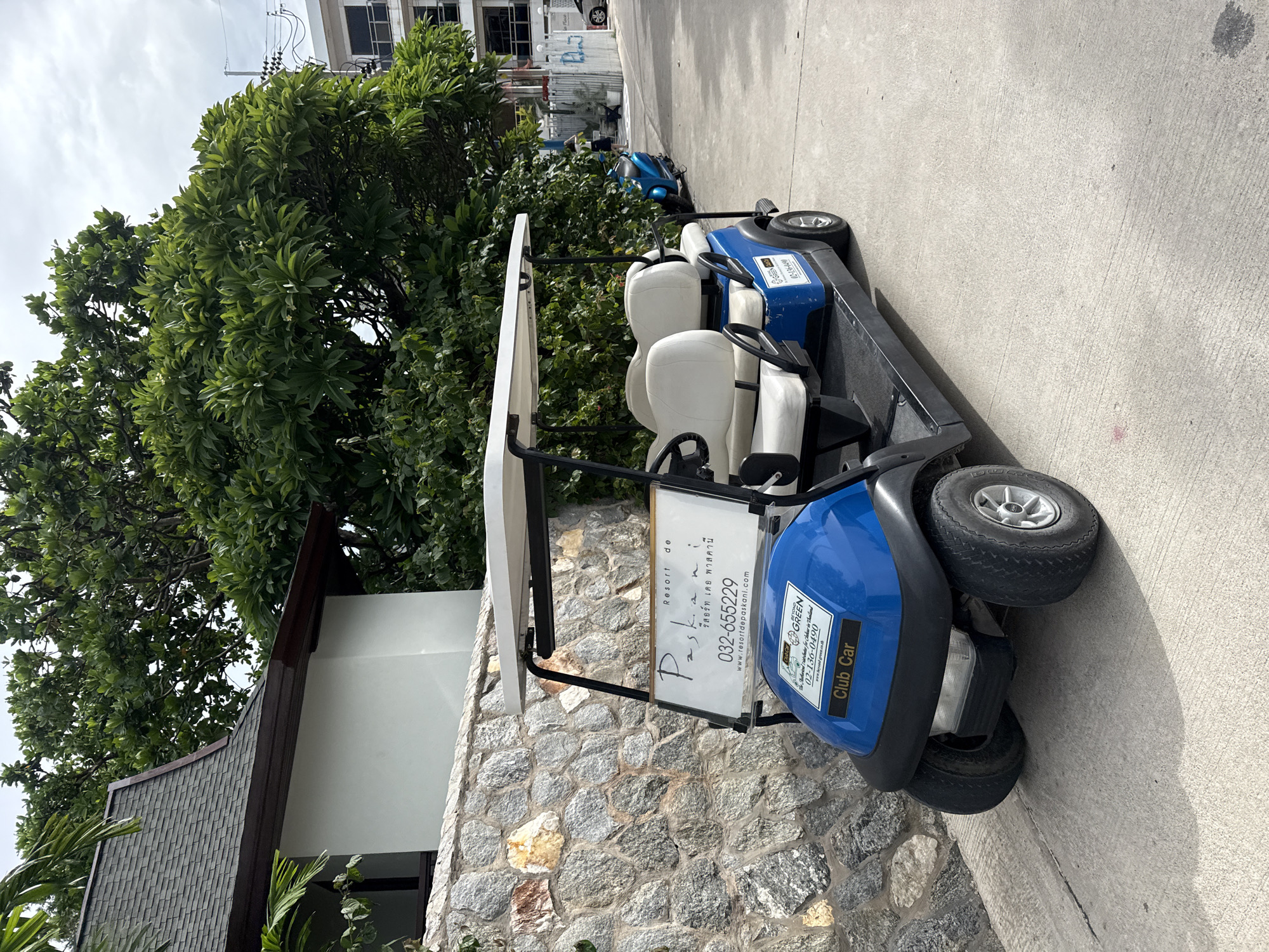 A blue and white four-seater golf cart is parked on a concrete path in front of a stone wall and a white building with a dark-tiled roof. The golf cart has the words "Resort de Paskani" and a phone number printed on a sign. A smaller sticker on the golf cart reads "BEYOND GREEN" with a phone number and website. The background shows lush green trees and a modern building with balconies. A blue scooter is parked further down the path, and a white car is visible in the distance. The sky is overcast. The location is implied to be within the Hua Hin City Municipality, Thailand, likely on the grounds of the Resort de Paskani.