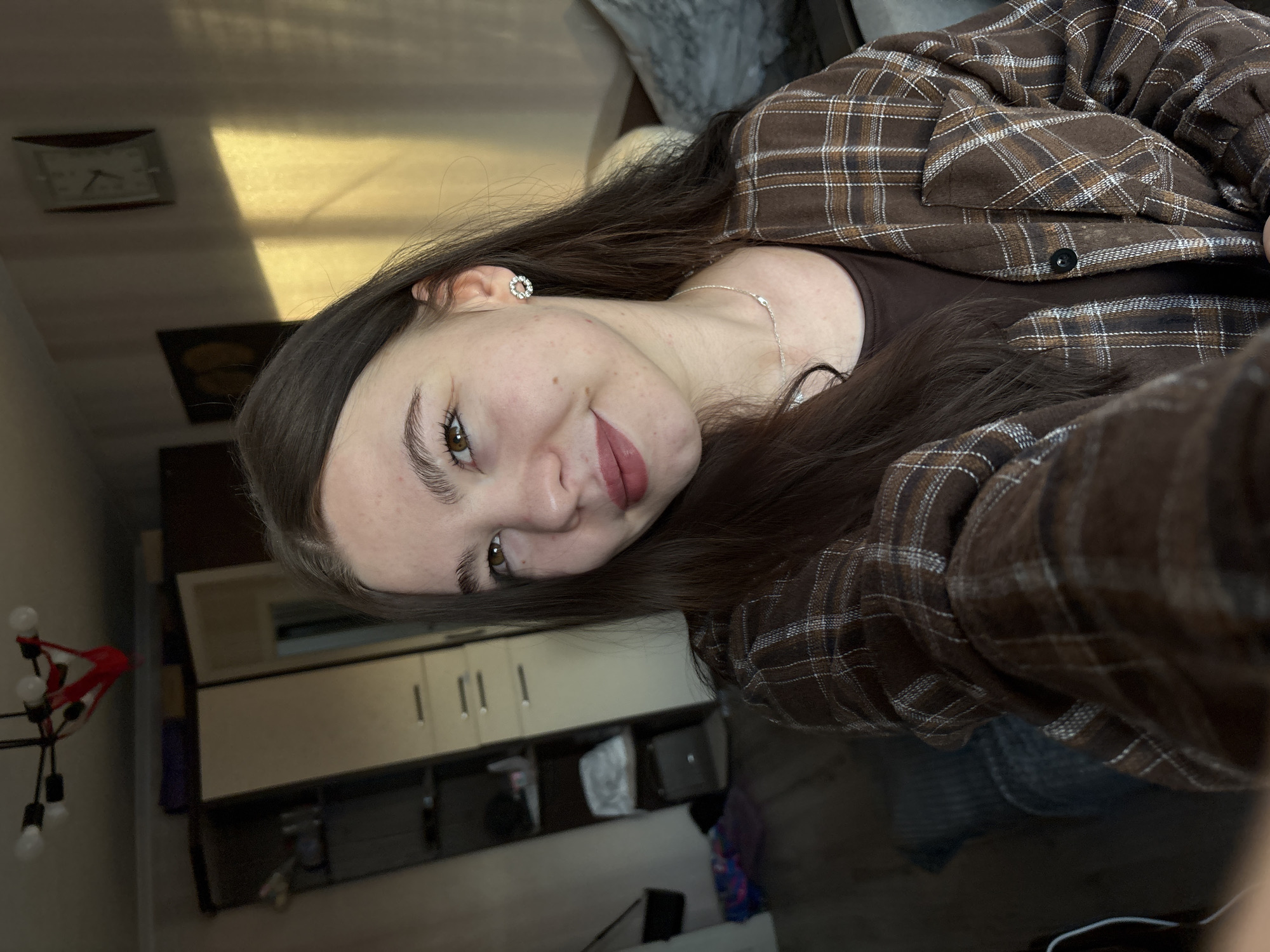 A photograph shows a young woman with brown hair, wearing a brown plaid shirt and a brown top. She is looking at the camera and smiling slightly. She has brown eyes, light skin, and wears earrings and a necklace. In the background, there is a wall with a clock, a dark artwork, and cabinets. There is also a lamp with red and white light bulbs on the left side of the frame. A blurry object in the background looks like a chair or sofa. The location is Perm, Russia.