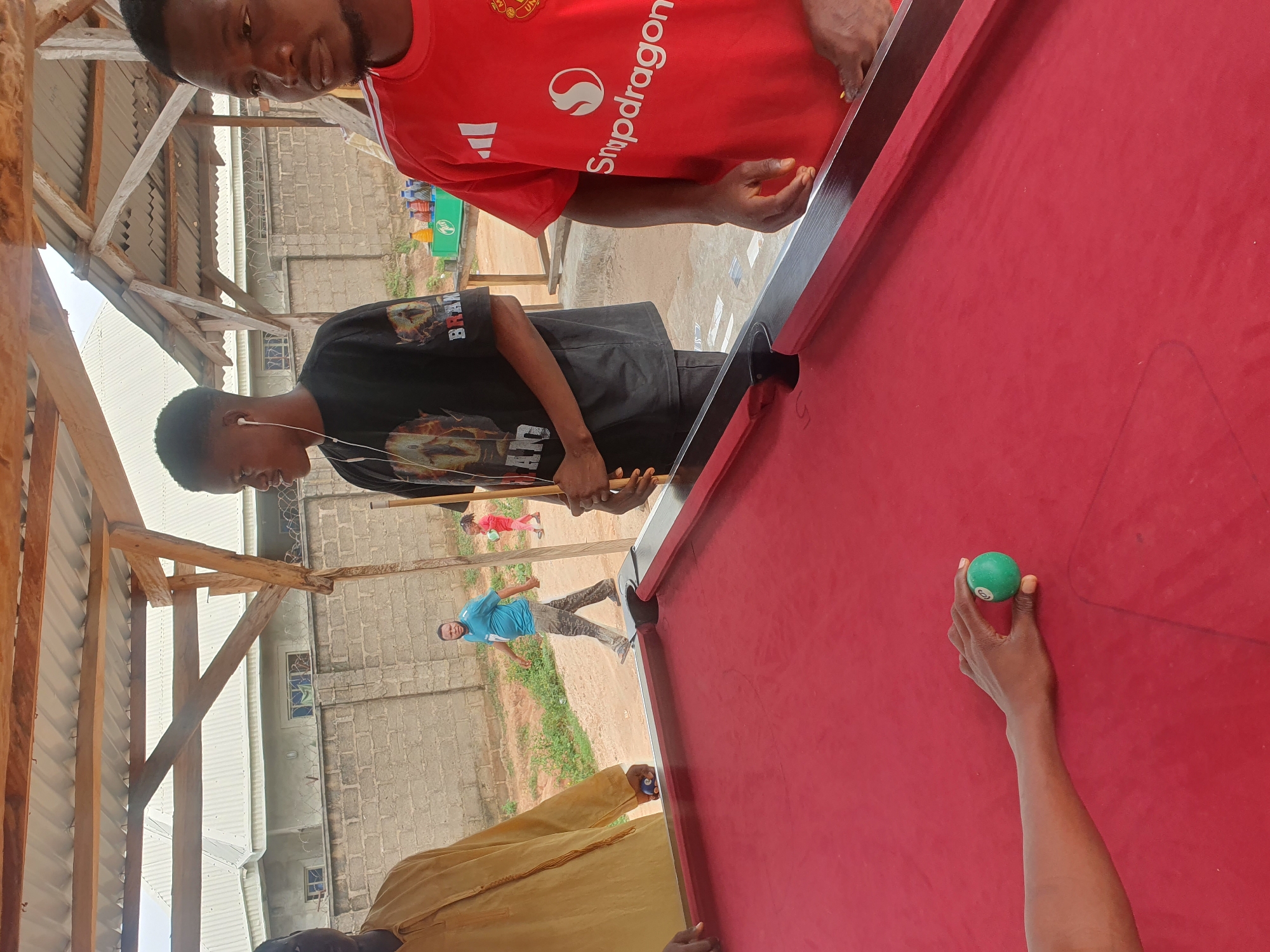 In Lokoja, Nigeria, at an outdoor billiard table with a red felt surface, a group of individuals are engaged in a game. The table is positioned under a rustic wooden roof structure, with corrugated metal sheeting overhead. A person on the left, partially visible, wears a yellow garment and holds a blue billiard ball. To their right, a young man in a black t-shirt with a graphic print and white earphones is preparing to play, holding a pool cue. Another young man to the right is wearing a red Manchester United jersey with "Snapdragon" branding. In the background, a man in a blue t-shirt walks away from the table, and a young child in a red outfit can be seen in the distance. The environment appears to be a casual outdoor setting with a brick wall and vegetation visible behind the billiard area.