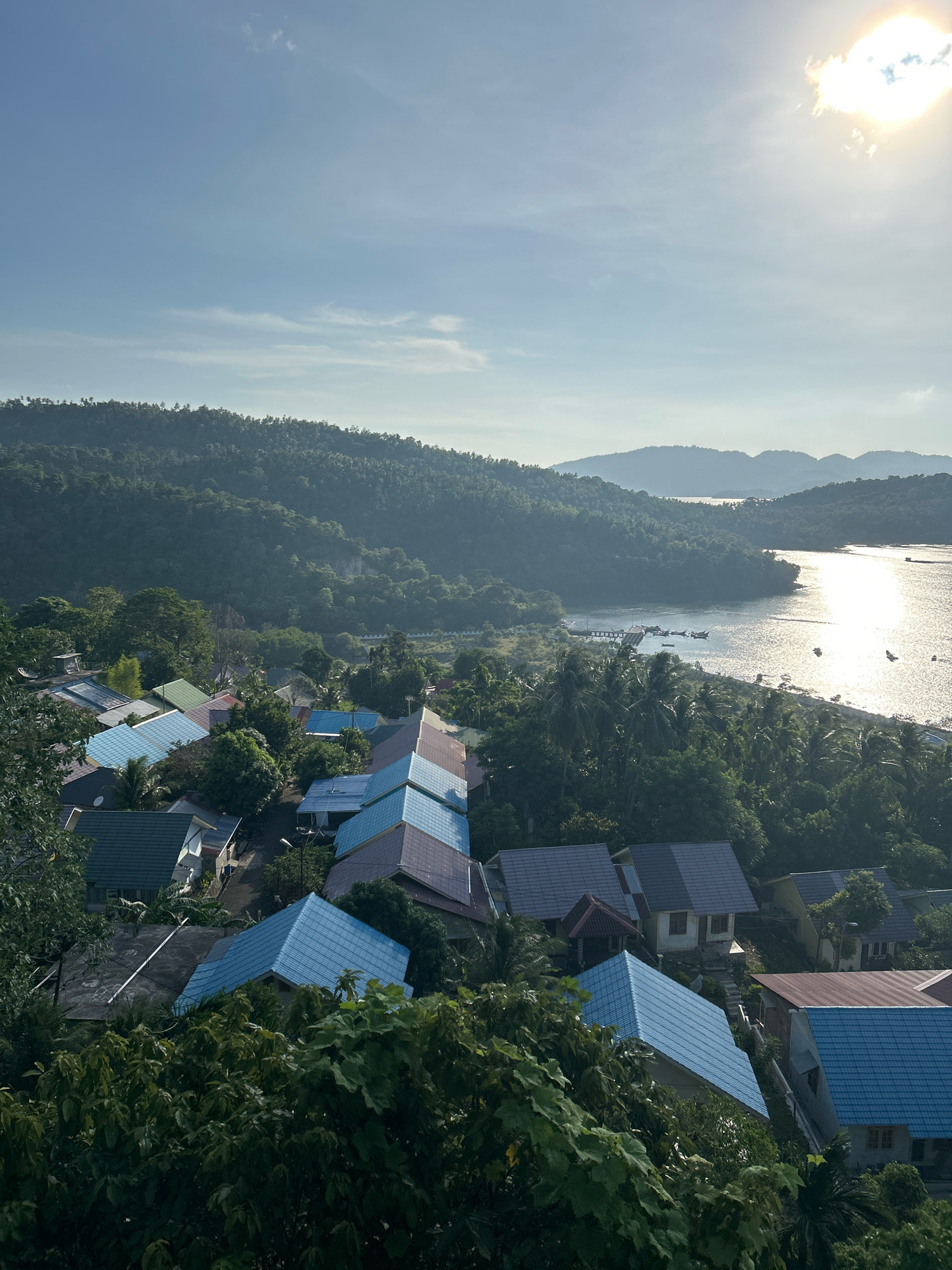 This aerial view captures a village nestled on a hillside overlooking a bay on the island of Weh, near Sabang, Indonesia. The foreground is dominated by lush green trees, obscuring some of the residential structures. A cluster of houses with colorful roofs, predominantly blue and brown, are visible on the slope. The village appears to be situated along a road that winds through the houses.
Beyond the village, a body of water glitters under the bright sun, reflecting the light from the sky. Hills covered in dense vegetation form the backdrop, with layers of mist or haze softening their outlines. Several boats are scattered across the water, and a small pier extends into the bay. The sky is a clear blue, with a few wispy clouds, and the sun is positioned in the upper right corner, casting a bright glow. The scene suggests a tranquil, tropical coastal environment.