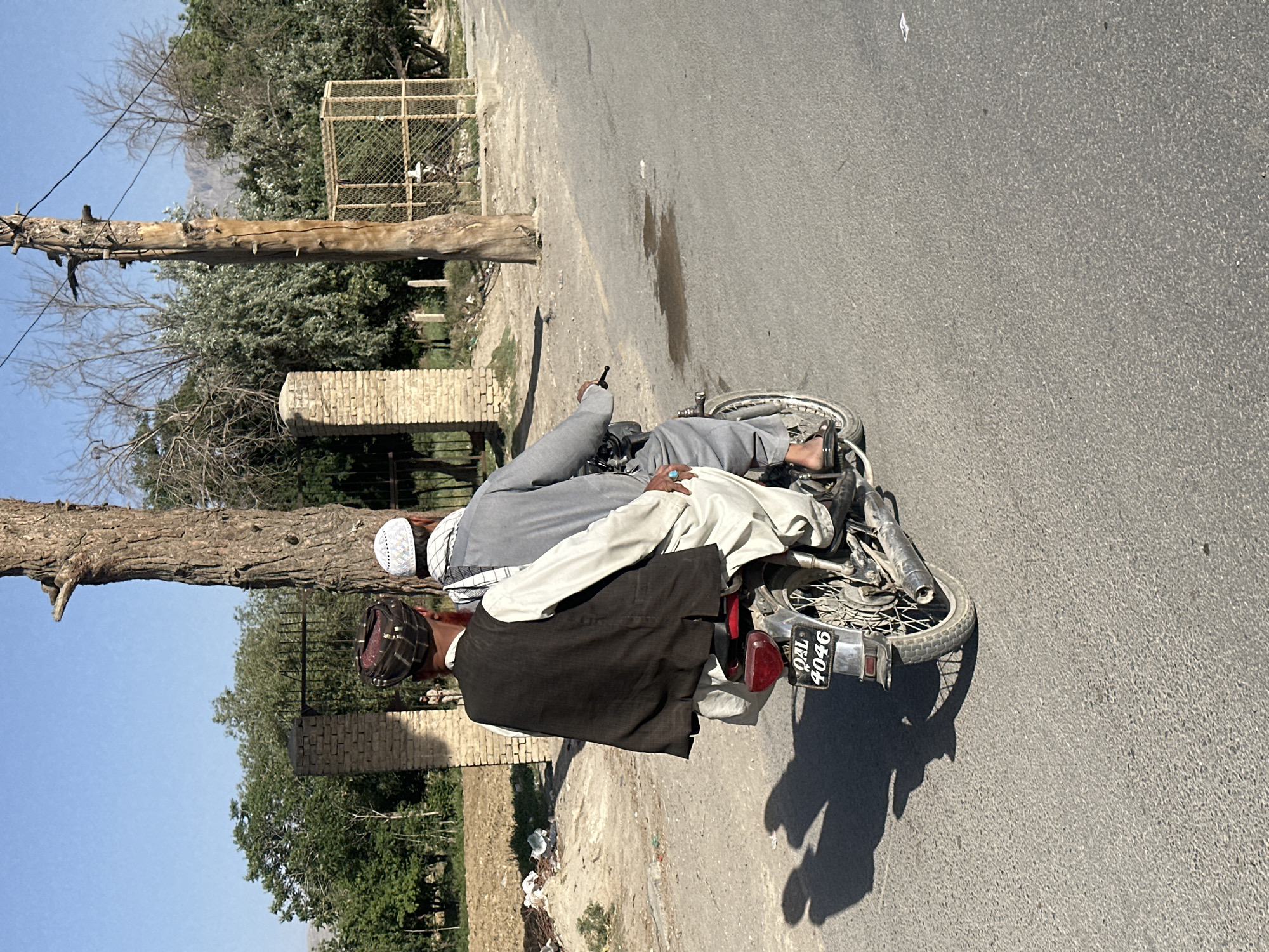 The image, when viewed upright, captures a scene on an asphalt road next to a dry, unpaved shoulder, under a clear blue sky. Two men are featured with a motorcycle that appears to be on its side.
One man, presumably the driver, is seen from behind, wearing a dark vest over light-colored clothing and a patterned dark head covering. He is positioned near the front of the motorcycle. The second man, a passenger, is lying across the motorcycle's seat and fuel tank, his body mostly visible from the back. He wears light grey attire, likely a shalwar kameez, and a white prayer cap (koofi). A notable detail is a turquoise ring on his right hand. His posture suggests he might be injured or unwell, being transported in an unconventional manner, or has possibly just fallen with the bike. His right leg is bent, and a bare foot in a sandal is visible.
The motorcycle itself is a typical model, lying on its left side. The rear wheel, exhaust pipe, and a black license plate are clearly visible. The text on the license plate reads "QAL 4046".
In the background, there's a mix of trees, some with green foliage and others appearing bare. A large, robust tree trunk or wooden pole stands upright on the left side of the scene. Further back, a low brick wall and a wire-caged structure are discernible. Faint outlines of mountains can be seen in the distance.
The long shadows cast across the road by the men and the motorcycle indicate either early morning or late afternoon. The weather appears sunny and clear. The overall impression is an incident or a stopped journey, possibly involving an injured individual.