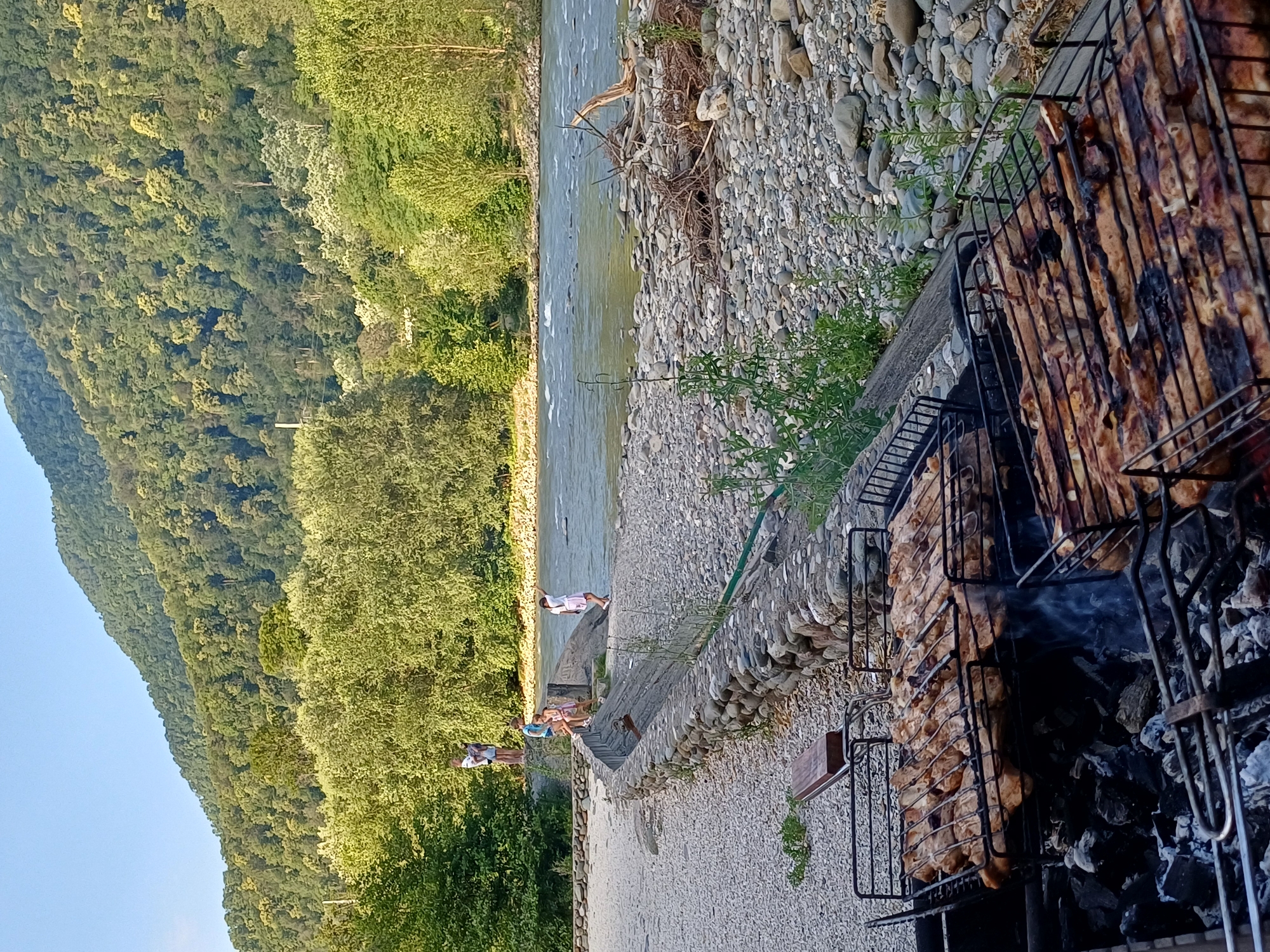 In Monastyr, Russia, a scenic view unfolds along a riverbank, with a focus on an outdoor barbecue. In the foreground, a charcoal grill is actively cooking several racks of meat, evident by the smoke rising and the browning of the food. Behind the grill, a pebbled embankment leads to a stone wall, bordering a grassy area. Further back, a serene river flows, with a forested mountain rising majestically in the distance, its slopes covered in lush green trees. A few individuals, including children, are visible near the river, enjoying the natural landscape.