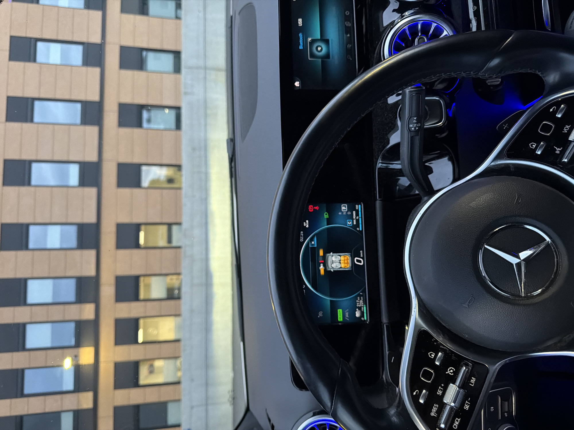 The image shows the interior of a car, looking out at a modern building with multiple rows of windows. The car's steering wheel and dashboard displays are in the foreground. The display on the left shows a car icon with battery charge indicators and the current speed as 0 km/h. The display on the right shows "Bluetooth USB" and an icon. A gear selector is visible on the right side of the steering column. Blue ambient lighting is present in the car's interior. The building in the background appears to be a hotel or office building. The overall scene suggests the car is parked. The question about Harstad, Norway is not supported by any visual information in the image.