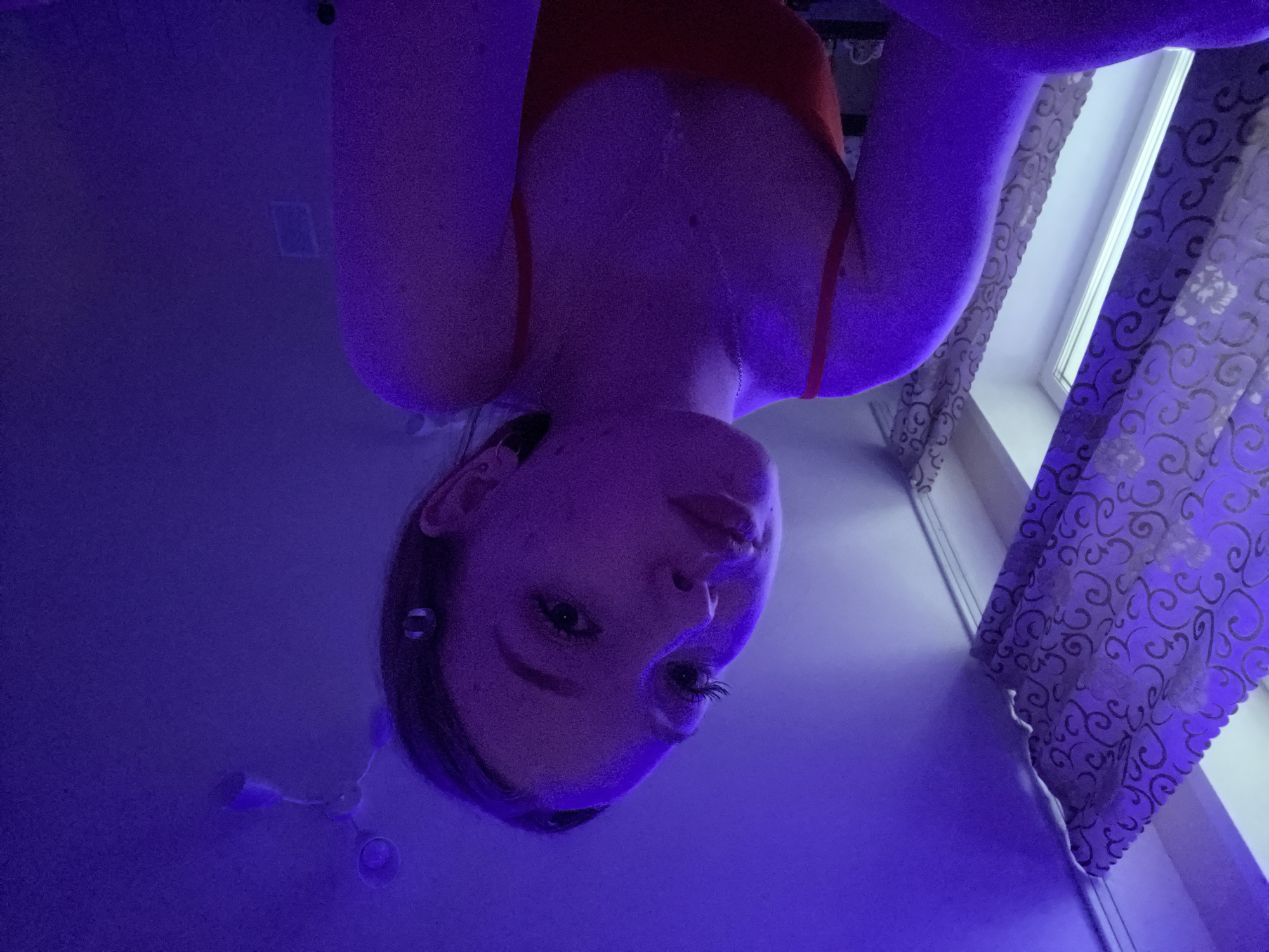 A selfie of a woman with long dark hair, fair skin and a reddish top, shot from a low angle looking up. The background includes a curtained window and an overhead light fixture. The overall lighting has a strong purple hue. The location is undeterminable from the image.