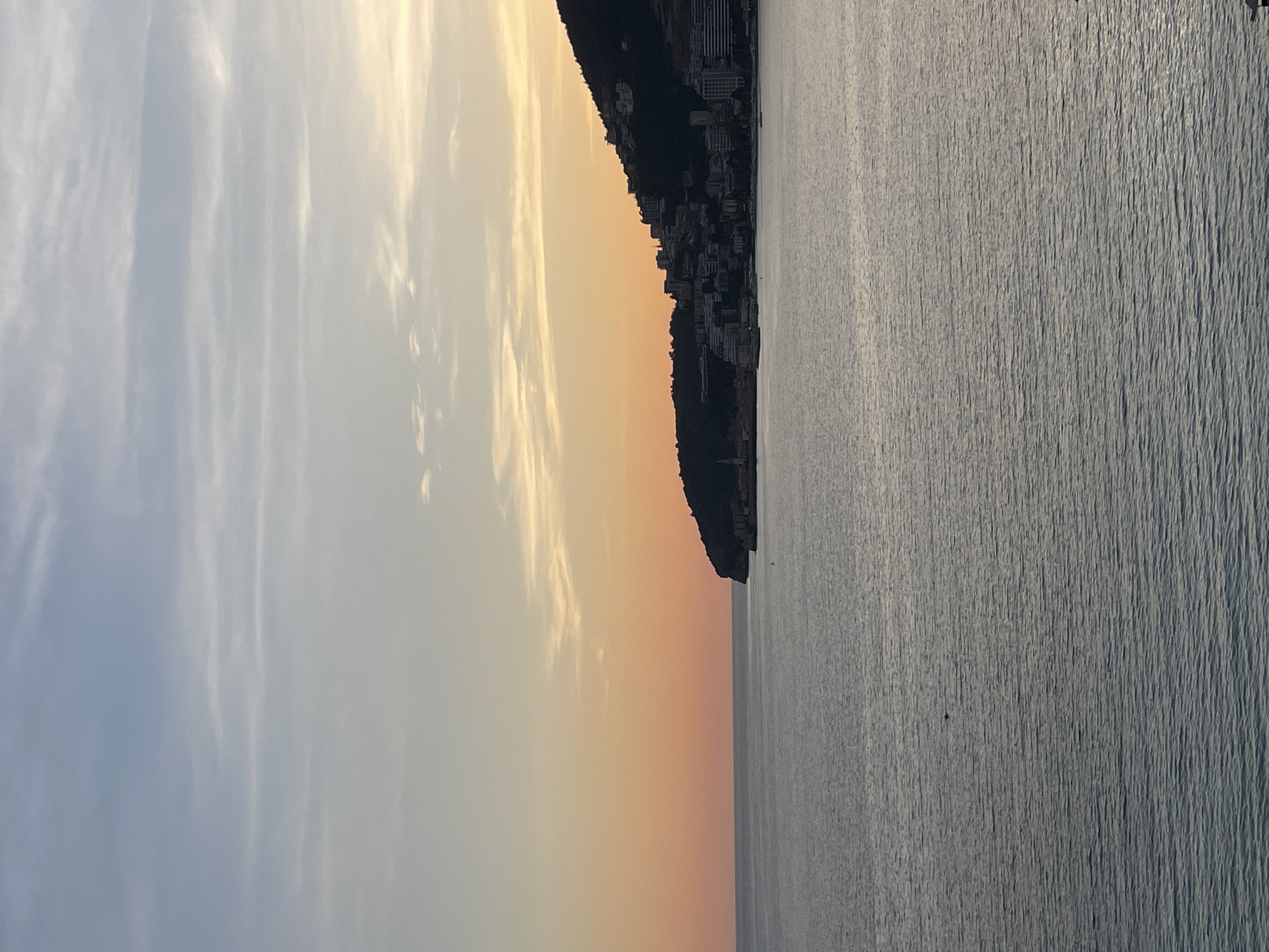 A wide shot captured at dusk or dawn depicts the coastal city of Budva, Montenegro, nestled against a hillside bordering the Adriatic Sea. The sky transitions horizontally from light blue with wispy clouds at the top to a warm gradient of orange and yellow near the horizon, indicative of a low sun angle. The lower half of the frame is dominated by the calm, dark blue-grey waters of the sea, textured by small, uniform ripples. A small, dark object, consistent with a buoy, is visible on the water's surface in the mid-left quadrant.
On the right side, a dark, tree-covered hill slopes down to the coastline. Along the base and lower slopes of this hill, the city of Budva is visible, characterized by numerous multi-story buildings and structures. The historic Old Town of Budva is identifiable as a fortified area on a small peninsula extending into the sea, featuring a prominent church bell tower with a pointed spire. Modern buildings, likely hotels or residential complexes, with light-colored facades and balconies, are distributed along the waterfront and ascending the hillside. No individuals or specific actions are discernible within the frame.
