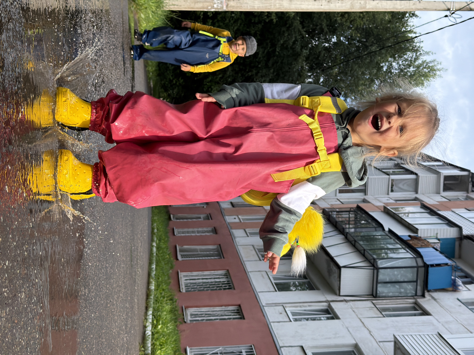 A young girl with blonde hair is jumping in a puddle, splashing water. She is wearing a pink rain suit over a green hoodie and yellow backpack with a fluffy yellow attachment. Her yellow boots are creating the splash. Behind her, a younger boy in a grey hat and blue rain pants over a yellow hoodie walks towards the viewer. They are on an asphalt surface with a large puddle in the foreground. In the background, there is a multi-story apartment building with balconies and windows, and some greenery. The scene appears to be in a residential area in the city of Chaikovsky, Russia, possibly on a cloudy day given the lighting.