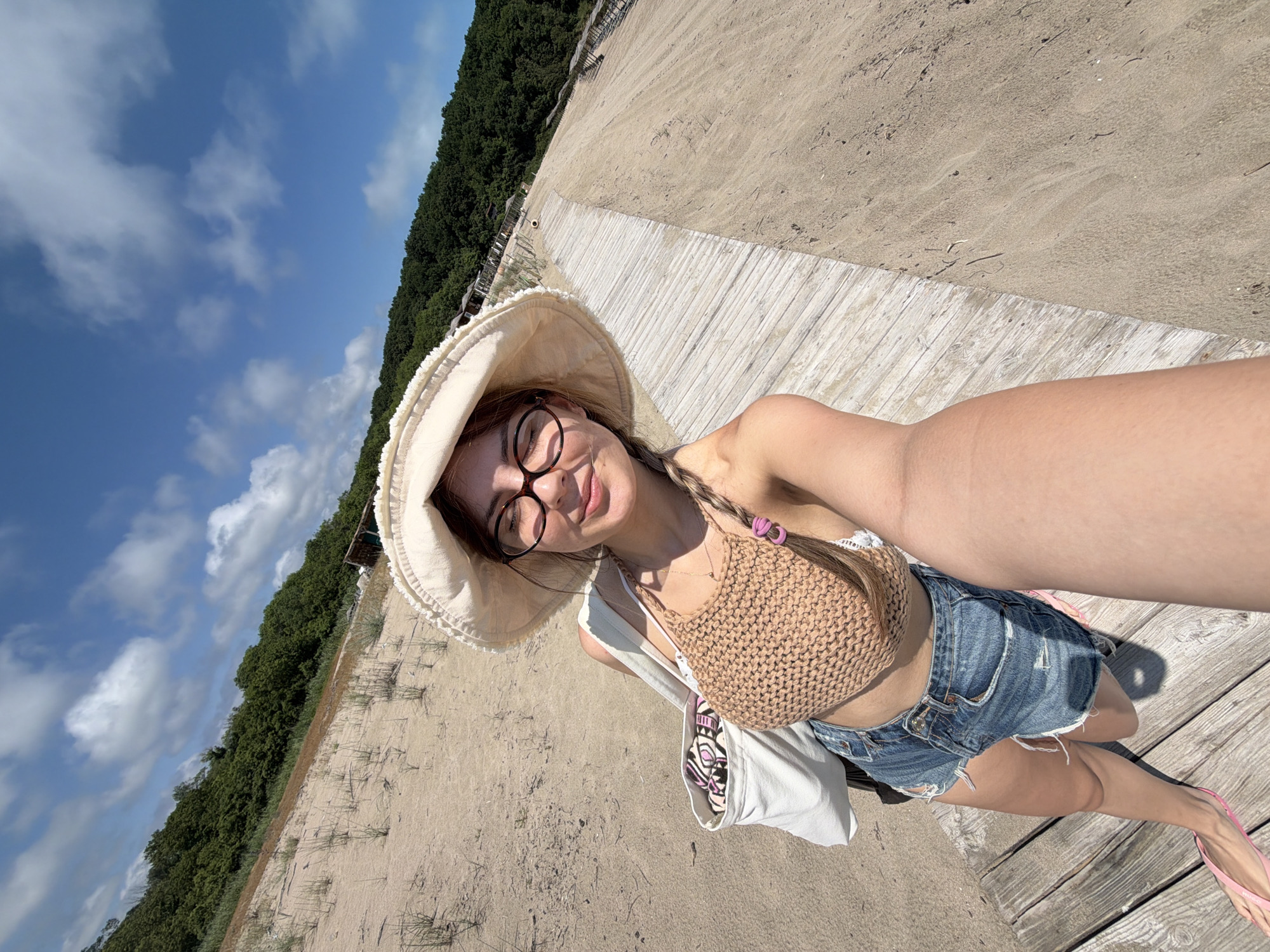 A young woman with dark hair is taking a selfie on a beach in Rezovo, Bulgaria. She is wearing a wide-brimmed sun hat, glasses, a crocheted bikini top, and denim shorts. She has a white tote bag slung over her shoulder. The beach is sandy with sparse vegetation, and a wooden boardwalk runs along the sand. In the background, there are trees lining a hill and a clear blue sky with scattered clouds. The sun is shining, casting shadows on the sand and boardwalk.