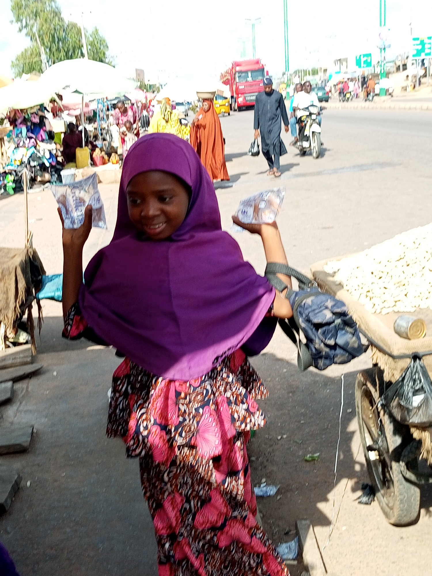 In Tempure, Nigeria, a young girl wearing a purple hijab and a colorful patterned dress is shown in the foreground, holding small packages in both hands. She appears to be smiling. Behind her, a market scene unfolds with stalls, merchandise, and other people. Some individuals are dressed in traditional attire, including a woman in an orange abaya carrying a basket on her head. In the background, a red truck and a motorcycle are visible on a road, indicating a busy urban environment. The ground is paved, and there are various objects scattered around, such as bags and wooden planks. The overall scene depicts a lively marketplace.