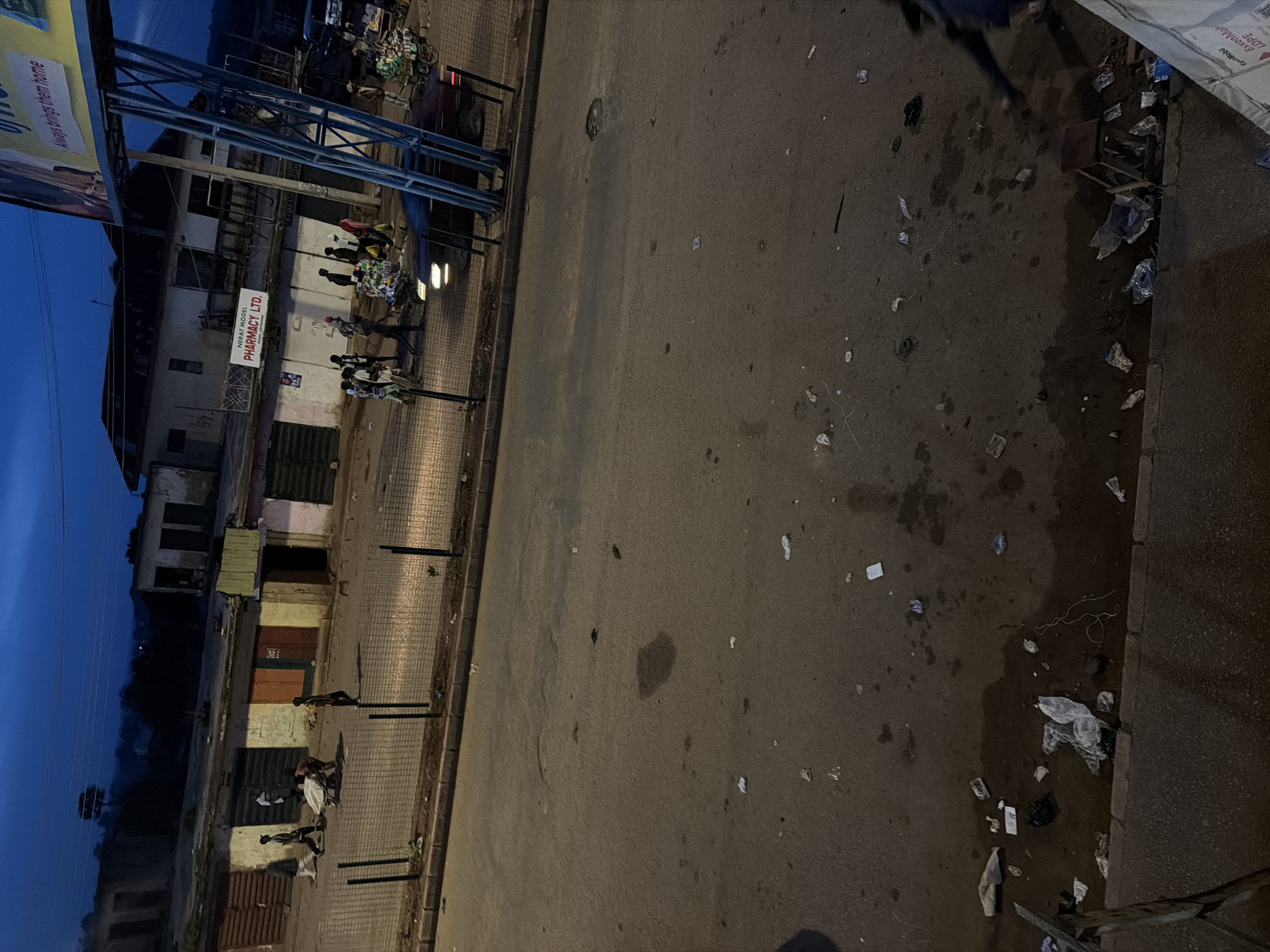 The image shows a street scene in Bukuru, Nigeria, at dusk. A road occupies the foreground, littered with various pieces of trash. In the middle ground, a low-rise commercial building complex runs parallel to the street. Several shop fronts are visible, some with closed shutters. A fence separates the sidewalk from the street. A few people are present in the middle ground, some walking and others near a wheelbarrow. A dark-colored car is visible on the street in front of the shops. In the background, more buildings and trees are visible under a dark blue sky. A large billboard with text is visible on the right side of the image.