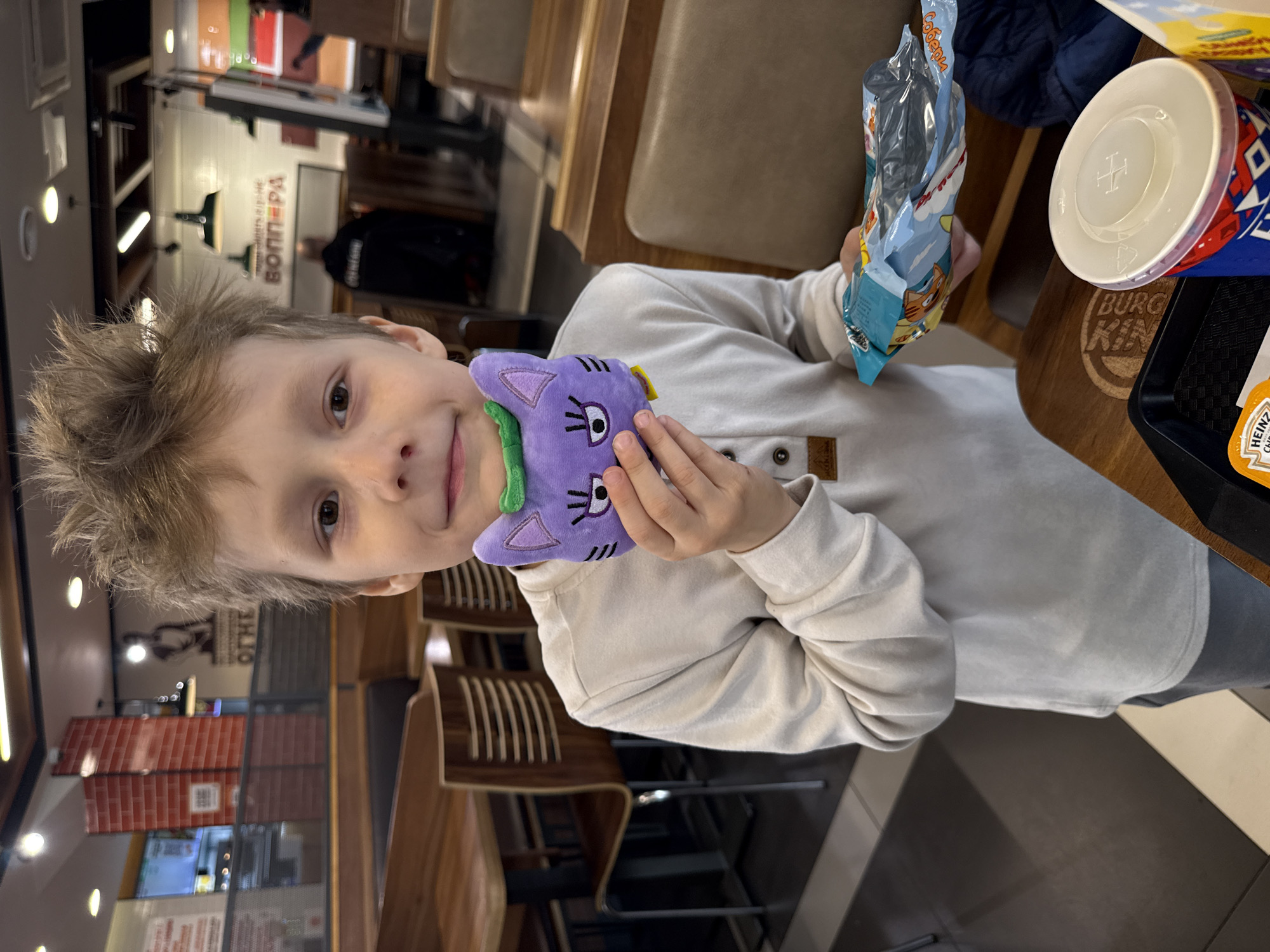 A young boy with short, light brown hair is seated at a table in what appears to be a fast-food restaurant. He is wearing a light-colored long-sleeved shirt with buttons at the collar. The boy is looking towards the camera with a slight smile.
In his left hand, he holds a small, purple plush toy that resembles a cat or monster, featuring large eyes, a pink nose, and green accents. His right hand holds an open, blue and white plastic snack bag, with some contents visible inside, possibly chips or a similar treat.
On the wooden table to his right, there's a red, white, and blue drink cup with a clear plastic lid, bearing a "BURGER KING" logo. Next to it, a black plastic tray holds a yellow "HEINZ KETCHUP" packet. The table itself also has the "BURGER KING" logo impressed into the wood.
The background shows the interior of the restaurant with wooden tables and chairs. Signs and posters are visible on the walls. One prominent sign features the word "ВОППЕР" (Vopper, or Whopper in Cyrillic) and another sign above it appears to say "ОТКРЫТО" (Open). Brick-patterned wall sections are also visible. In the deeper background, another person, blurry and wearing a dark jacket, can be seen sitting. The lighting suggests an indoor setting, likely during the day or evening. The boy appears content and engaged with his toy and snack. The snack bag also has the word "Собери" (Collect) printed on it.