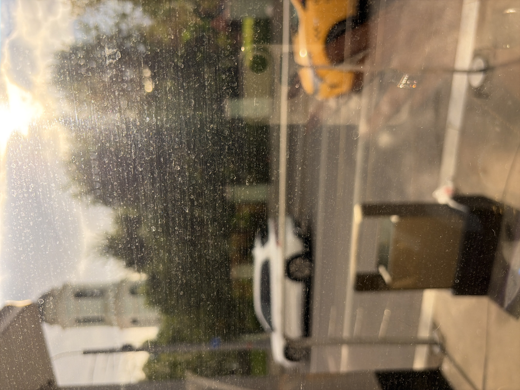 The image shows a view through a dirty window, likely from a ground-floor establishment, overlooking a street scene in Moscow, Russia. The sunlight is bright, suggesting it is daytime.
In the background, a tall, white bell tower with a green roof and spire is visible, partially obscured by trees. This architectural feature suggests the location is near a church or monastery. A white SUV is driving on the street in front of the trees and the bell tower. To the right, a yellow taxi is parked or stopped.
In the foreground, through the dirty glass, a dark, metallic trash receptacle with a brown insert is visible. The glass itself is covered in numerous smudges, water spots, and streaks, indicating it has not been cleaned recently. The overall impression is of a typical urban street scene viewed from an interior space.