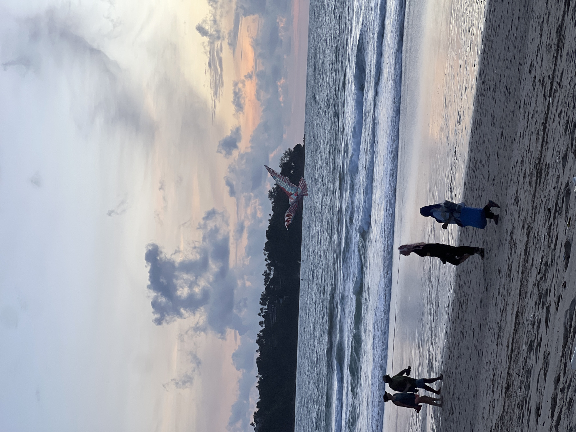 The image depicts a beach scene at dusk in Choeng Thale, Thailand. A kite in the shape of a bird, with red, white, and blue patterned wings, is flying over the ocean. The ocean has moderate waves and a distant shoreline with trees and buildings is visible. The sky is filled with pastel-colored clouds. On the sandy beach, several individuals are walking. In the foreground, two people, a woman wearing a headscarf and a light blue outer garment, and a person in a dark garment with pink hair, are walking towards the right. Further to the left, two other figures, one wearing a dark top and shorts, and another in a green jacket and shorts, are walking towards the left. The sand is wet in places, reflecting the sky's colors.