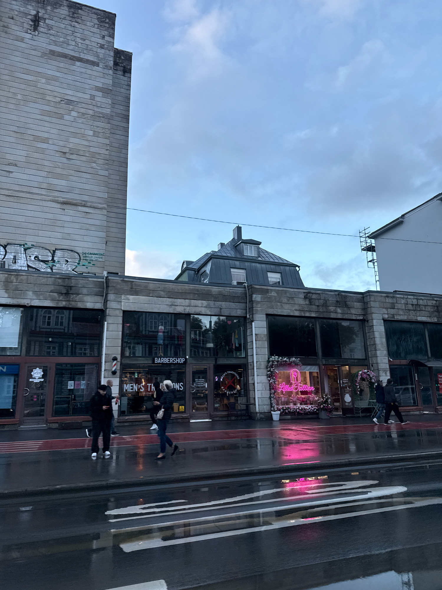 The image shows a street scene in Tallinn, Estonia, on a cloudy day. The street is wet, indicating recent rain, and reflections of lights and buildings are visible on the asphalt. On the right side of the image, there is a building with several shopfronts. One shop has a neon sign that reads "Lillafallin" and is decorated with flowers. Another shop displays a sign for "BARBERSHOP" and "MEN'S TRADITIONS". There are a few people walking on the sidewalk. The architecture features modern and older elements, with a large, plain stone-clad building on the left and a more detailed building with large windows and a mansard roof in the center. Graffiti is visible on the upper section of the stone-clad building.