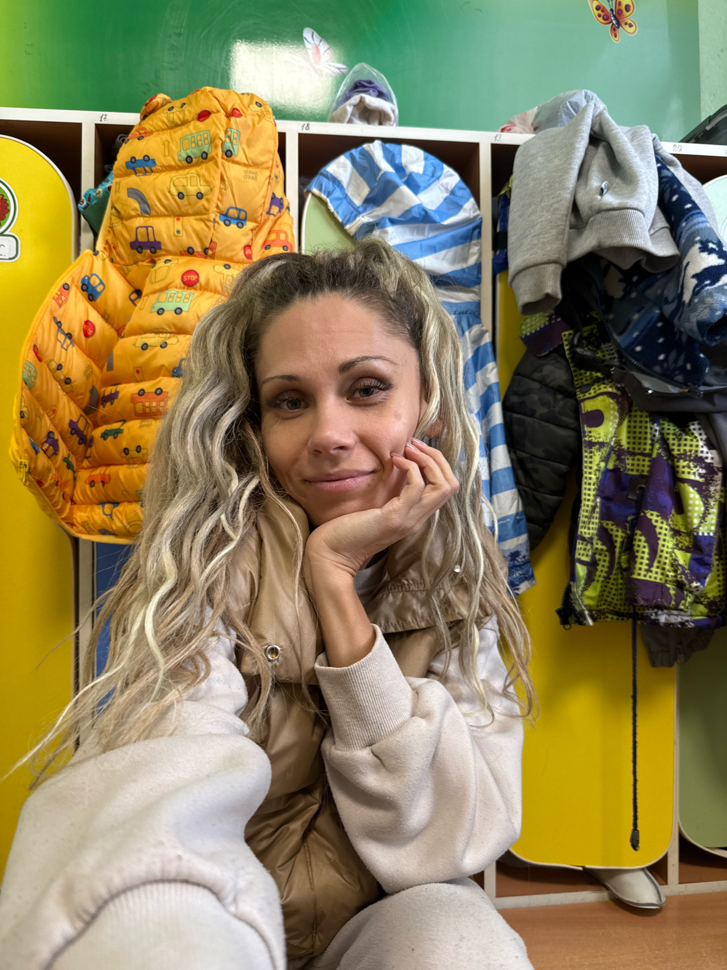 A woman with long, blonde, wavy hair is taking a selfie in what appears to be a children's room or daycare facility. She is wearing a tan puffer vest over a cream-colored sweatshirt. Her left hand is resting on her chin, and she is looking directly at the camera with a slight smile. Behind her, a row of colorful cubbies holds children's clothing, including a bright orange jacket with a car print and a blue and white striped jacket. The walls are painted green, and a butterfly sticker is visible on the upper left wall. The scene suggests a casual, personal moment captured in a child-oriented environment.