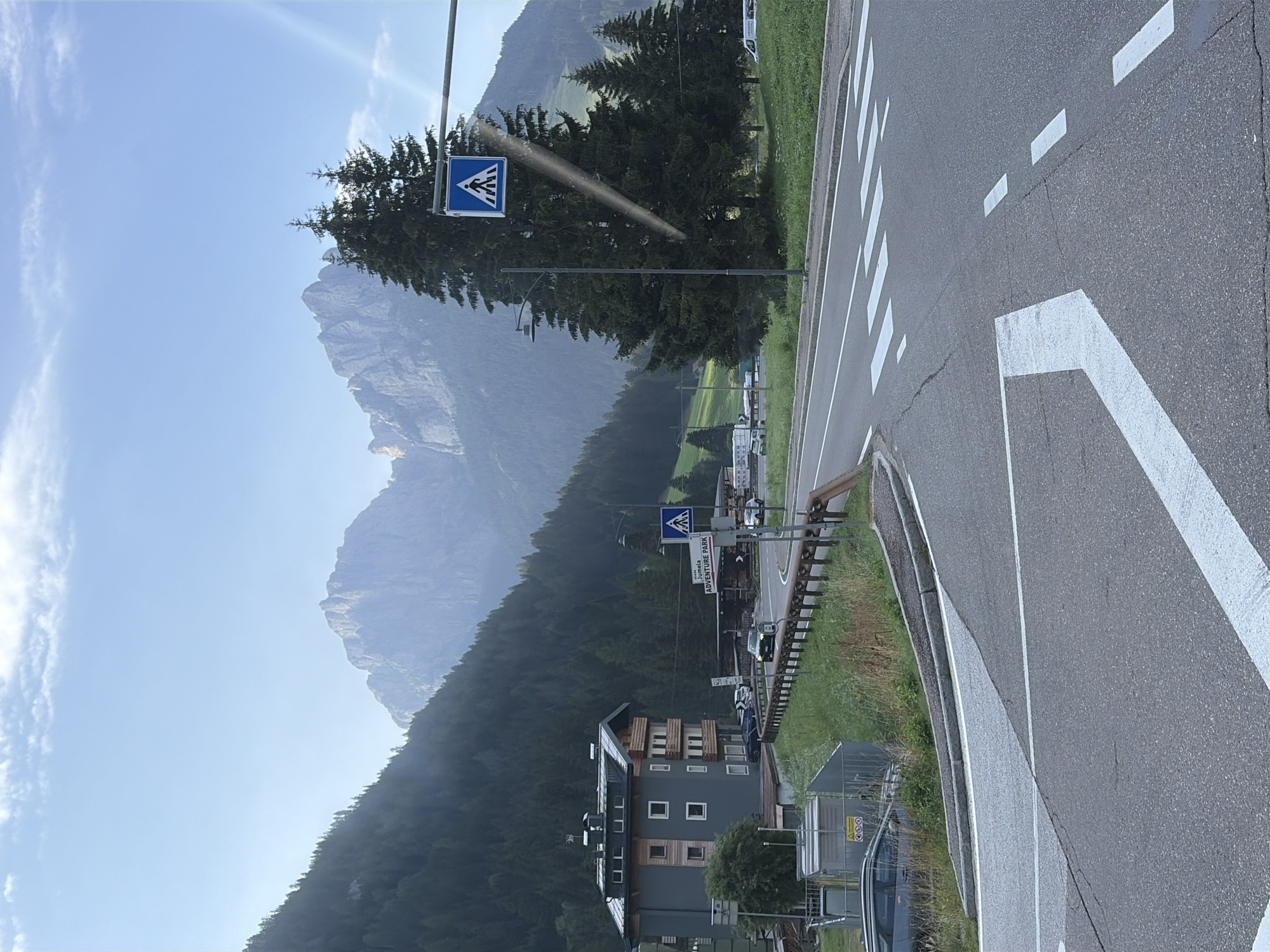 The image shows a road scene in the vicinity of Pera, Italy. In the foreground, a paved road with white lane markings and a crosswalk is visible, with a slight incline. To the left, a grey car is parked alongside a grassy embankment and a fence enclosing some electrical equipment. A modern, multi-story building with a grey facade and wooden accents is situated on a hillside to the left of the road. Behind the building, a dense forest covers a steep mountain slope.
In the middle ground, a two-lane road curves to the right. Several cars are present on the road, including a dark colored vehicle driving towards the viewer and a white van parked in the distance. A pedestrian crossing sign with a blue background and a white pictogram of a person walking is mounted on a pole, to the right of the road. Further to the right, a cluster of tall evergreen trees stands against a backdrop of rugged, mist-shrouded mountains. A street lamp is also visible near the pedestrian crossing sign. The sky above is a pale blue with scattered clouds. A signpost on the right side of the road indicates "Strada JUMETA ADVENTURE PARK".