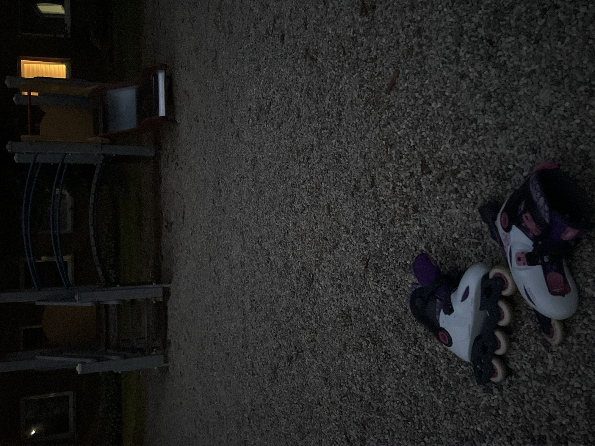 The image depicts a dark outdoor scene, likely a playground, with a gravel ground surface. In the foreground, two pairs of rollerblades are lying on the gravel. The rollerblades are white with purple and pink accents, and have visible wheels. In the background, a yellow and red playground structure with a slide is visible. The structure appears to be made of metal and wood. There are also windows of a building visible in the background, with one window emitting a bright yellow light. The lighting suggests it is nighttime or dusk. The image does not provide any specific visual cues to pinpoint a location within the city of Vilnius, Lithuania.