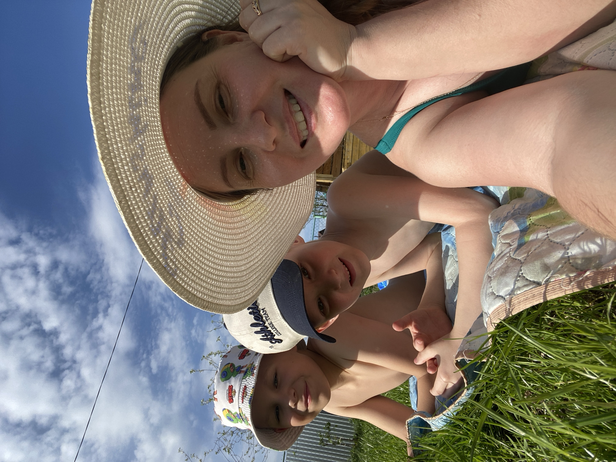 The image is a selfie taken from a low angle, showing a woman and two children outdoors, presumably on a sunny day. The location is Chaikovsky, Russia.
The woman is wearing a large straw hat and a green swimsuit, smiling at the camera. One of the children is wearing a baseball cap with the words "COLLEGE TEAM," and the other is wearing a bucket hat with colorful cartoon prints. Both children are also smiling.
They are sitting on a quilted blanket spread on green grass. In the background, there is a blue sky with white clouds, and glimpses of a metal fence and foliage can be seen.