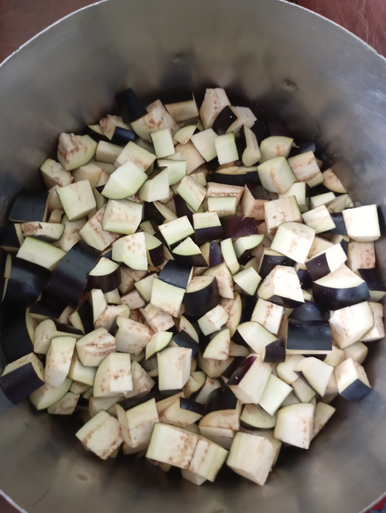 The image shows a close-up, top-down view of a large metal pot filled with diced eggplant. The eggplant pieces are cut into roughly uniform cubes, with some showing the dark purple skin and others the pale white flesh. The pieces are packed densely within the pot, suggesting they are prepared for cooking or storage. The setting appears to be a kitchen or food preparation area, indicated by the metal pot and the visible edge of what might be a countertop or table. There is no visible text in the image, and no people are present. The lighting suggests it is daytime, but there are no specific cues about the weather or exact time. The overall impression is one of domestic food preparation in Horishni Plavni, Ukraine.