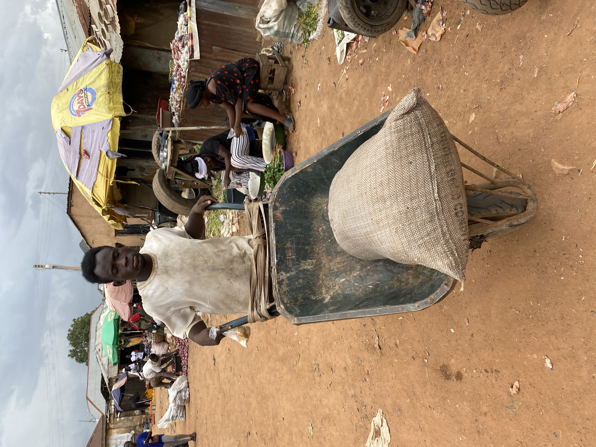 In Bukuru, Nigeria, a young Black man in a dirty white t-shirt pushes a wheelbarrow containing a large burlap sack. The wheelbarrow's handles are wrapped with a brown strap, and the man holds a small plastic bag in his right hand. In the background, a busy marketplace unfolds under a cloudy sky. Various vendors are visible, some tending to stalls piled with produce, while others engage in transactions. Structures with corrugated metal roofs and makeshift awnings are prominent. The ground is a dusty, reddish-brown dirt. Two women sit on a wooden stool and a low bench, respectively, appearing to be selling or preparing goods, with bowls of green vegetables in front of them. One woman wears a black dress with red heart patterns, and the other a white headscarf and striped pants. The overall atmosphere suggests a typical day of commerce in an outdoor market.