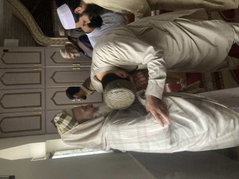 An indoor scene in Quetta City Tehsil, Pakistan, shows a group of approximately six adult males. In the foreground, a younger man, wearing a light tan shalwar kameez and a dark woven cap, is bowing forward, kissing the hand of an older man. The older man, with a long white beard, is wearing a light-colored, possibly plaid, shalwar kameez and has a checkered fabric wrapped around his head and shoulders. His right hand rests on the younger man's left arm. Both individuals are looking towards each other.
In the mid-ground, other men are visible. To the right of the central interaction, a man with a dark beard and a white prayer cap is partially visible, looking downwards. Behind the younger man, another man with a dark beard and dark clothing looks to the left. Further back, a man with a grey beard and a patterned cap is smiling.
The environment features light-colored wall cabinets with arched, decorative paneling and brass-colored trim. A window is visible on the far left. A patterned red and white rug is visible on the floor in the lower right corner, extending into the background. The lighting suggests an indoor setting, possibly a mosque or madrasa.