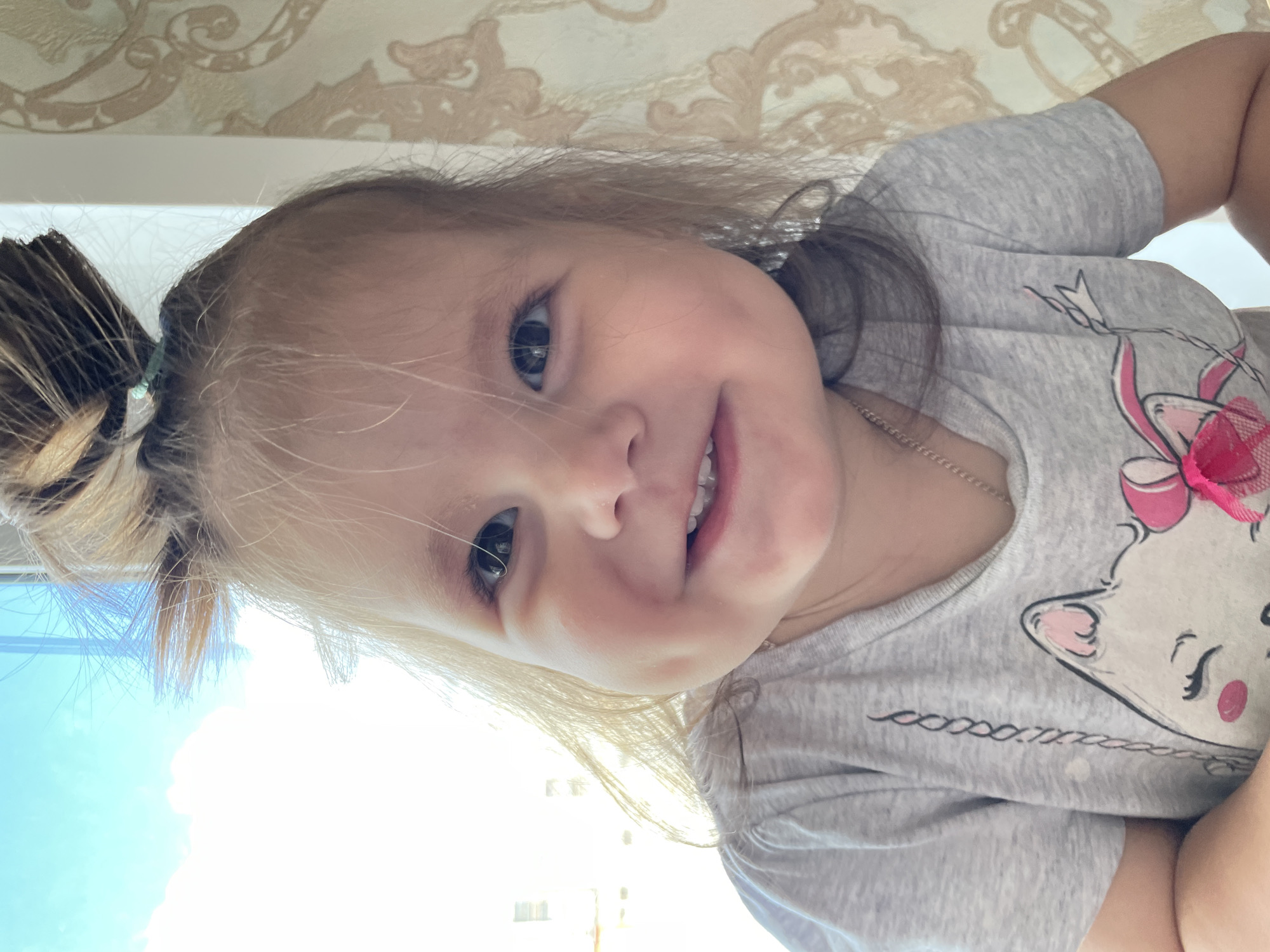 A brightly lit, close-up portrait shows a young child, likely a girl, with blonde hair pulled into a ponytail on the left side of her head. She looks directly at the viewer with wide, dark eyes and a joyful, open-mouthed smile, revealing her teeth. She wears a light grey t-shirt featuring a cartoon character, possibly a cat, adorned with a pink bow and details. A thin silver chain is visible around her neck.
The background is split between an indoor and outdoor view. Above the child's head, decorative wallpaper with a light brown and beige patterned motif is visible. To the left and below her, bright daylight streams in through a window, showing a clear, light blue sky, indicating it is daytime with good weather. The child's happy expression radiates warmth and cheerfulness.
On the grey t-shirt, below the cartoon character, a decorative design can be seen. It appears as a chain of small, intertwined 'o'-like shapes followed by some indistinct text or symbols, possibly "Xoxo T". The scene is likely set indoors, in a home environment in Izhevsk, Russia.