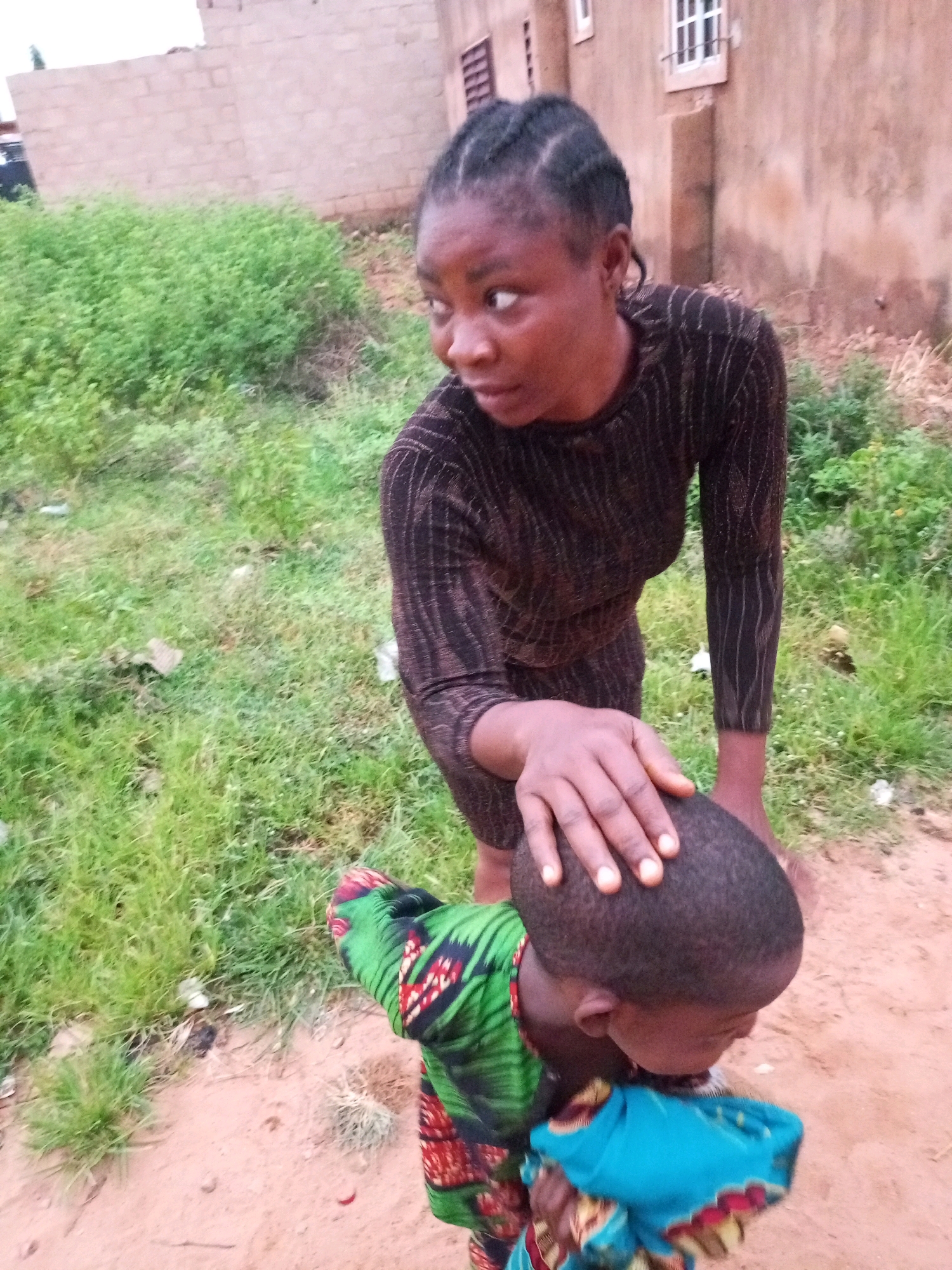 In Tempure, Nigeria, a woman with braided hair is bending over to place her hand on the head of a child who is facing away from the camera. The woman is wearing a brown, long-sleeved dress with a ribbed texture. The child is wrapped in a colorful fabric, predominantly green with red and black patterns, and a blue cloth is visible around their neck. They are standing on a dirt path, with dry grass and some green vegetation surrounding them. In the background, there is a building constructed with light-colored bricks, and a portion of another building with a window is also visible. The sky appears overcast.