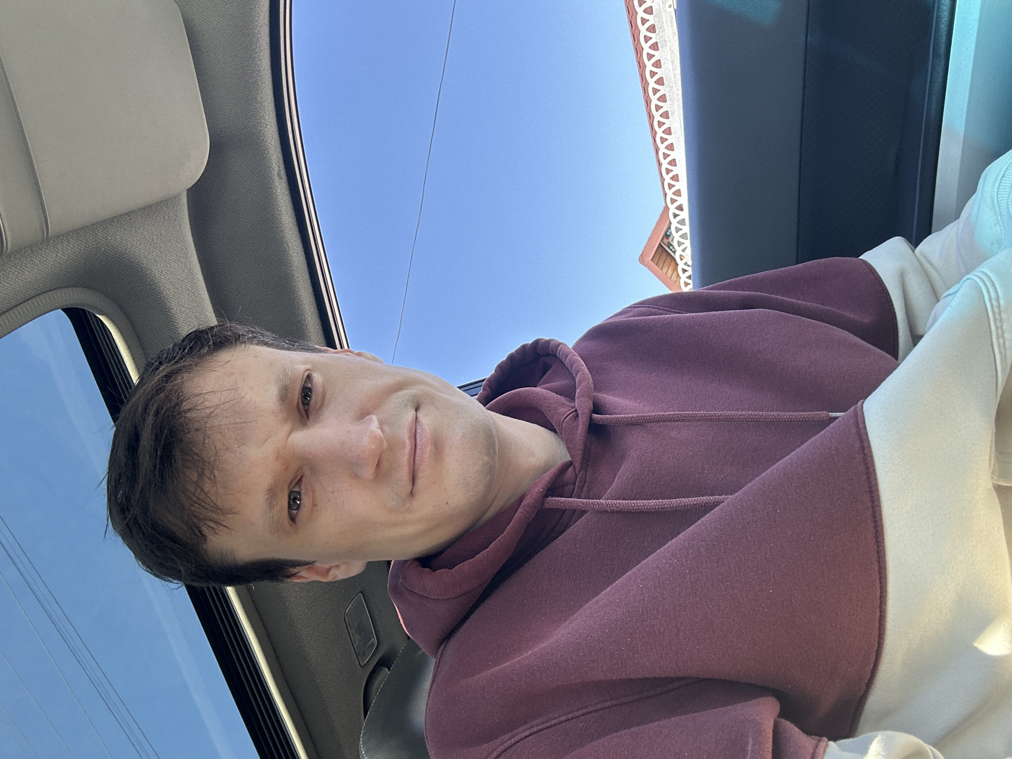 A man is pictured in a selfie, taken from inside a car. He is wearing a maroon and cream hooded sweatshirt. The car has a sunroof open, revealing a clear blue sky. In the background, through the car window, the roof of a building with distinctive white decorative trim is visible, along with a glimpse of trees. The scene appears to be in Makiv, Ukraine.