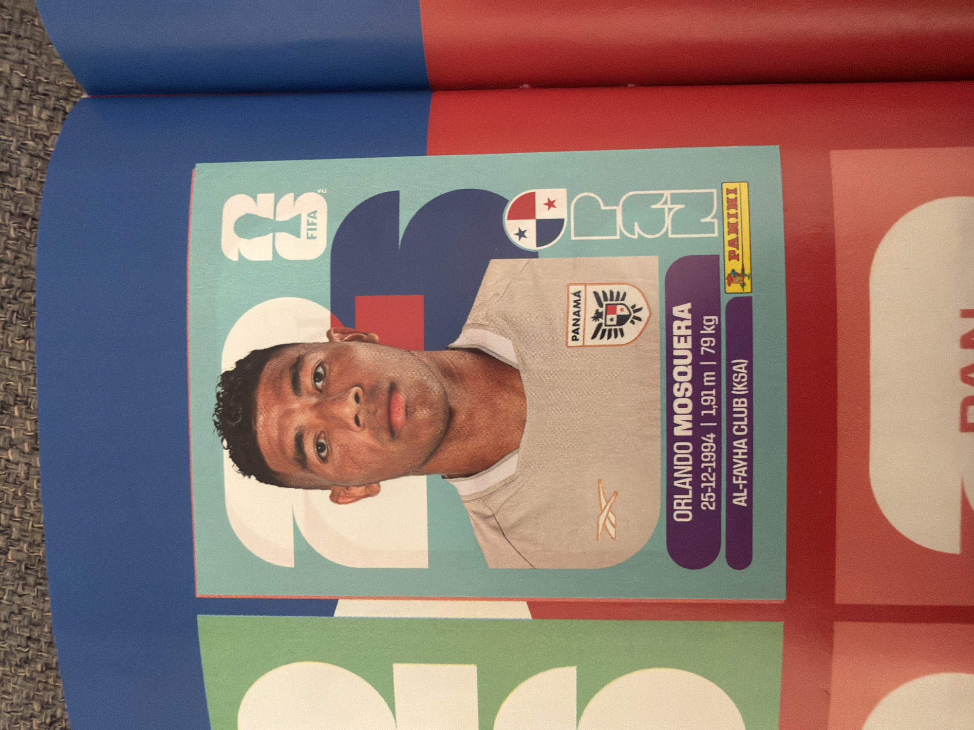 The image shows a close-up of a sports trading card featuring a soccer player. The player, identified as Orlando Mosquera, is shown from the chest up, looking directly at the camera. He is wearing a light-colored jersey with an orange logo on the left side of his chest. The card has a light blue background with large, abstract shapes and the FIFA logo. Below the player's image, his name, birthdate (25-12-1994), height (1.91 m), weight (79 kg), and club (Al-Fayha Club (KSA)) are listed. A small image of the Panamanian flag and a Panini logo are also present on the card. The card is placed within a larger publication, possibly a sticker album, as indicated by the surrounding blue and red borders and the texture of the pages. The background of the image shows a textured surface, possibly a table or a rug. There is no visible information or context that would suggest this image is related to the city of Zlatitrap, Bulgaria.