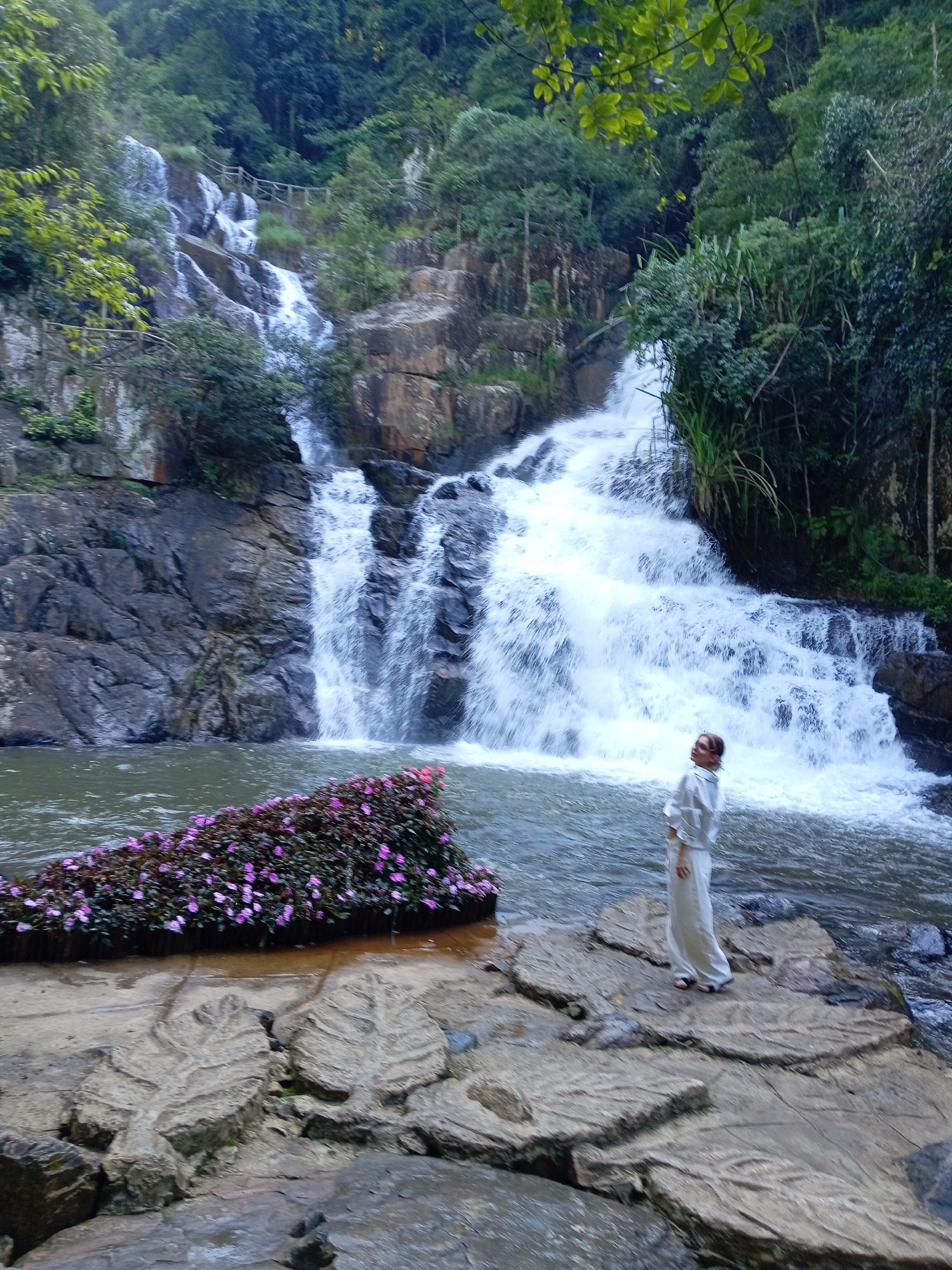 A woman in a white outfit stands on large, flat rocks near the base of a cascading waterfall in Da Lat, Vietnam. The waterfall flows down a rocky cliff face covered in lush green foliage. In the foreground, a cluster of vibrant pink flowers is arranged on a floating platform in the water. The woman is facing the waterfall, looking upwards, with her hands clasped in front of her. The water is a murky green, and the rocks are wet and dark. Trees and vegetation surround the waterfall, creating a natural and serene environment. The image appears to be taken from a slightly elevated position, looking down towards the waterfall and the woman.