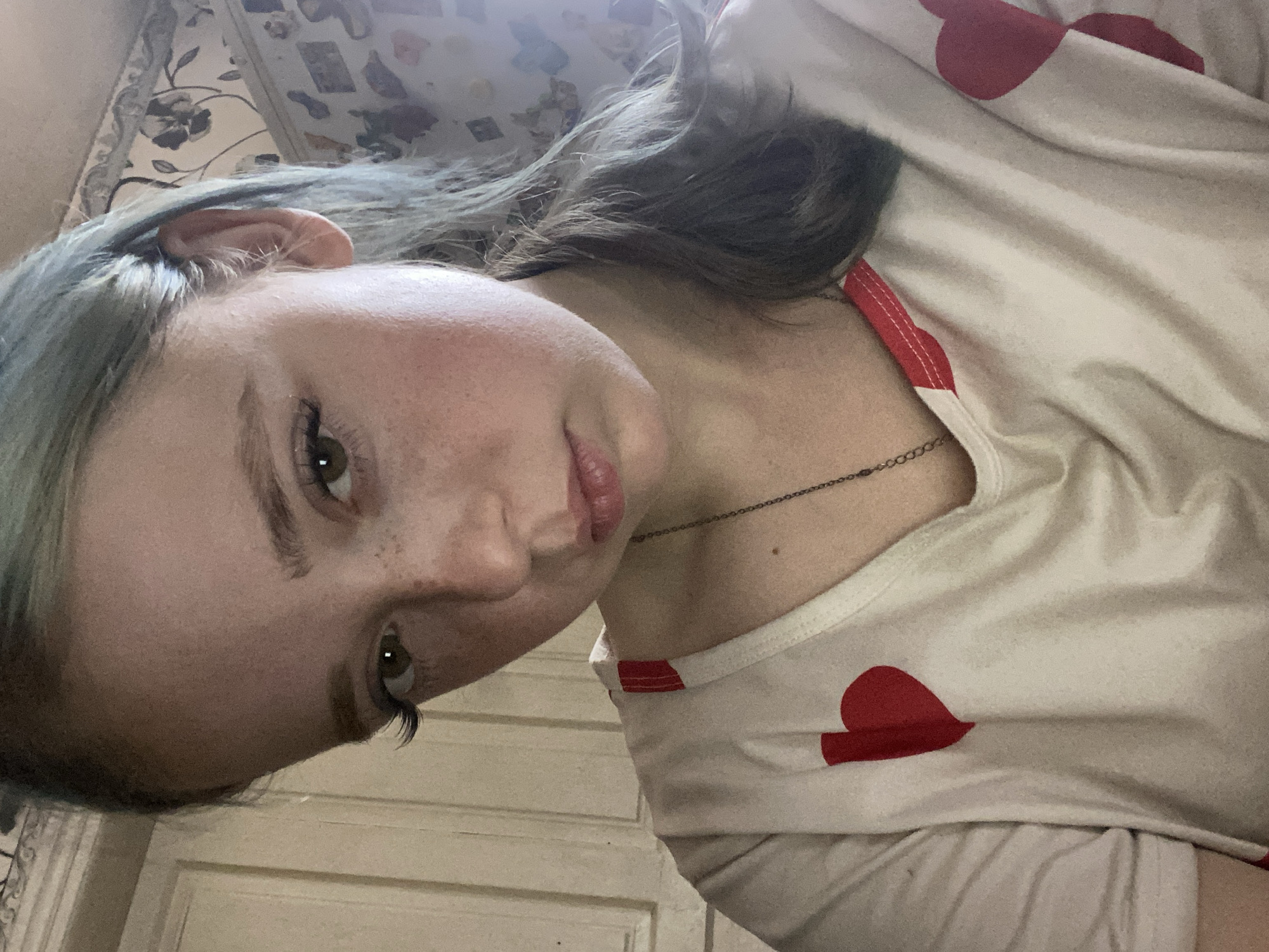 The media file is a close-up, low-angle selfie of a young person with light skin and freckles. They have light-colored hair with a greenish tint and expressive green eyes with dark eyelashes. The person is wearing a light-colored t-shirt with a pattern of red hearts. A thin chain necklace is visible around their neck. The background appears to be an indoor setting with a wall that has a wood-paneled texture and possibly a floral patterned tapestry or wallpaper. The lighting is soft and appears to be coming from above and slightly to the left, suggesting it might be daytime. The person's head is tilted, and their gaze is directed towards the camera, conveying a relaxed or introspective mood. There is no visible text in the image. The location context provided is Nova Praha, Ukraine.