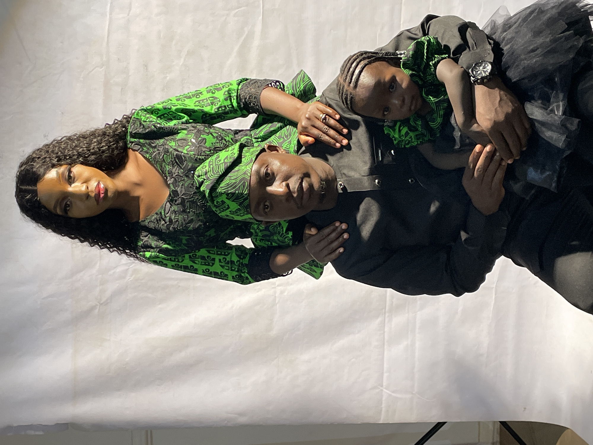 Three individuals are centrally featured, against a white backdrop. A woman with long, dark, curly hair is wearing a green and black patterned top. A man, wearing a black top and a green and black patterned head wrap, has a child resting on him. The child wears a green and black patterned top and a black tulle skirt. The man's right wrist is adorned with a watch. The location is suggested to be Jos, Nigeria.