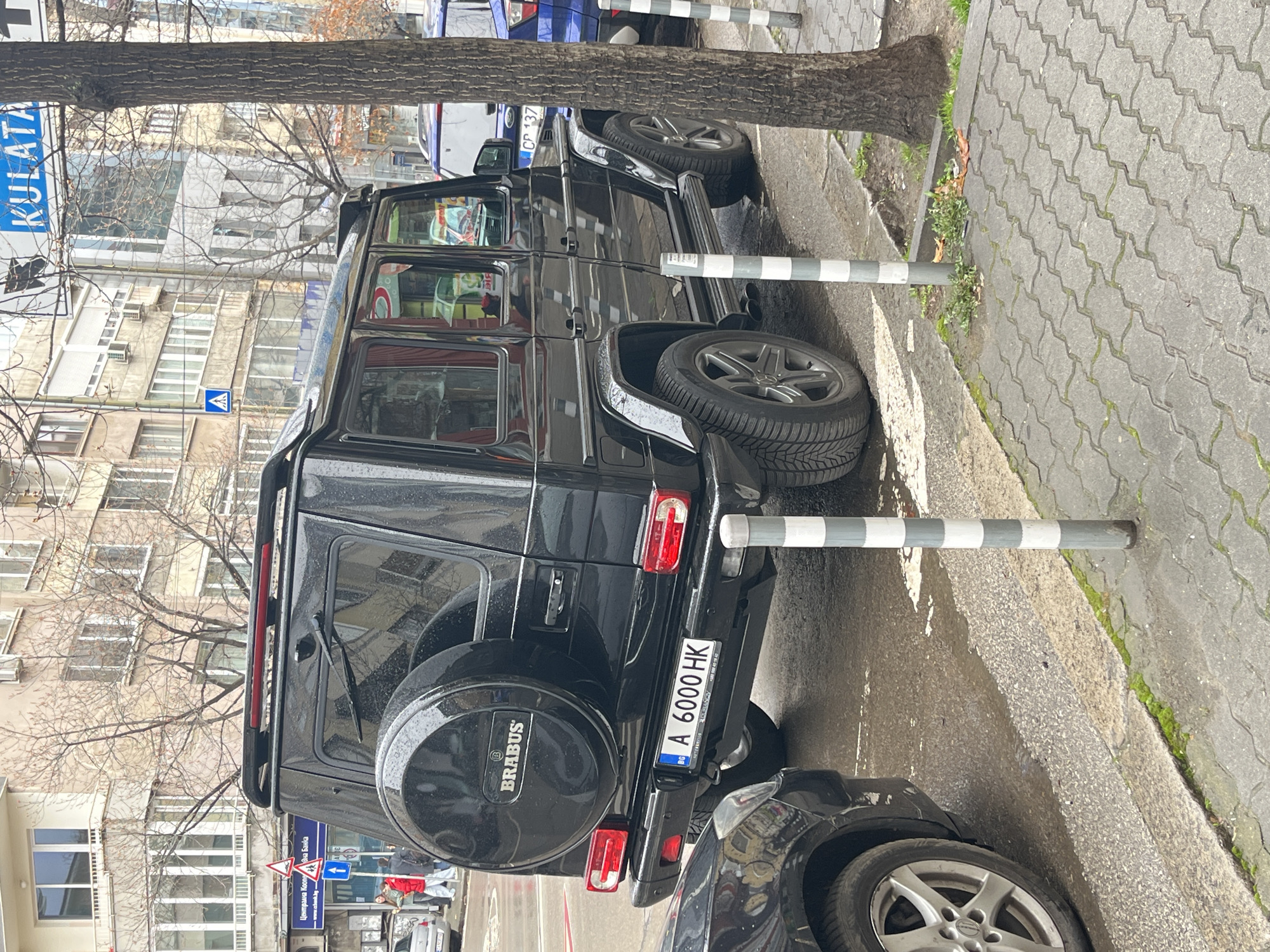 A black Brabus G-Wagon with Bulgarian license plate A 6000 HK is parked on a wet street in Sofia, Bulgaria. The car is positioned next to a tree and between two striped bollards. A second black car is partially visible behind the Brabus. A street sign with the text "KULATA" is visible in the background, along with buildings and bare trees. Pedestrians are also visible in the background.