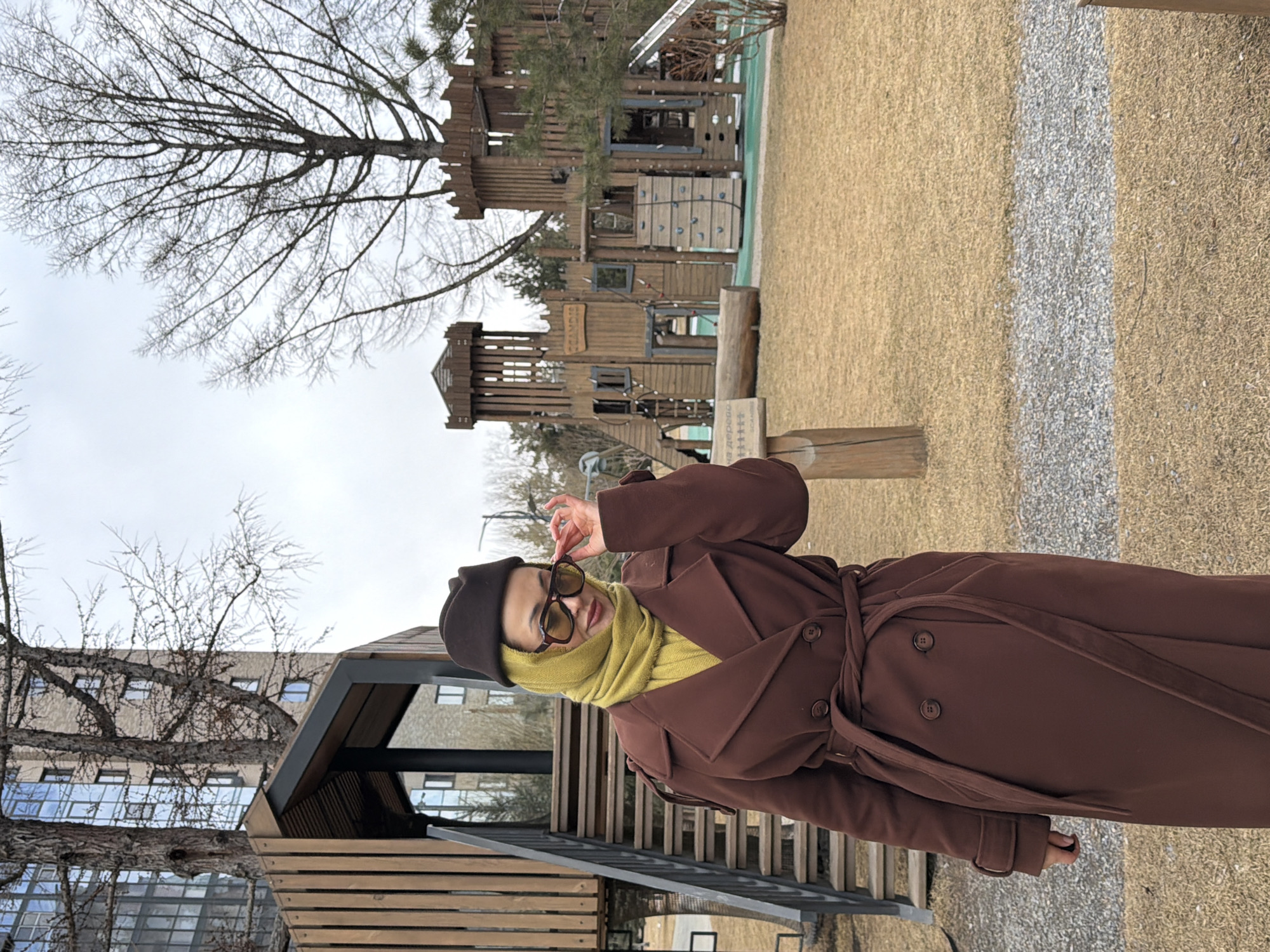 A woman wearing a brown coat, brown hat, yellow scarf, and sunglasses stands outside near a playground. The playground is made of wood and has multiple levels. A gravel path and trees are also visible. The location is Krasnoyarsk, Russia.