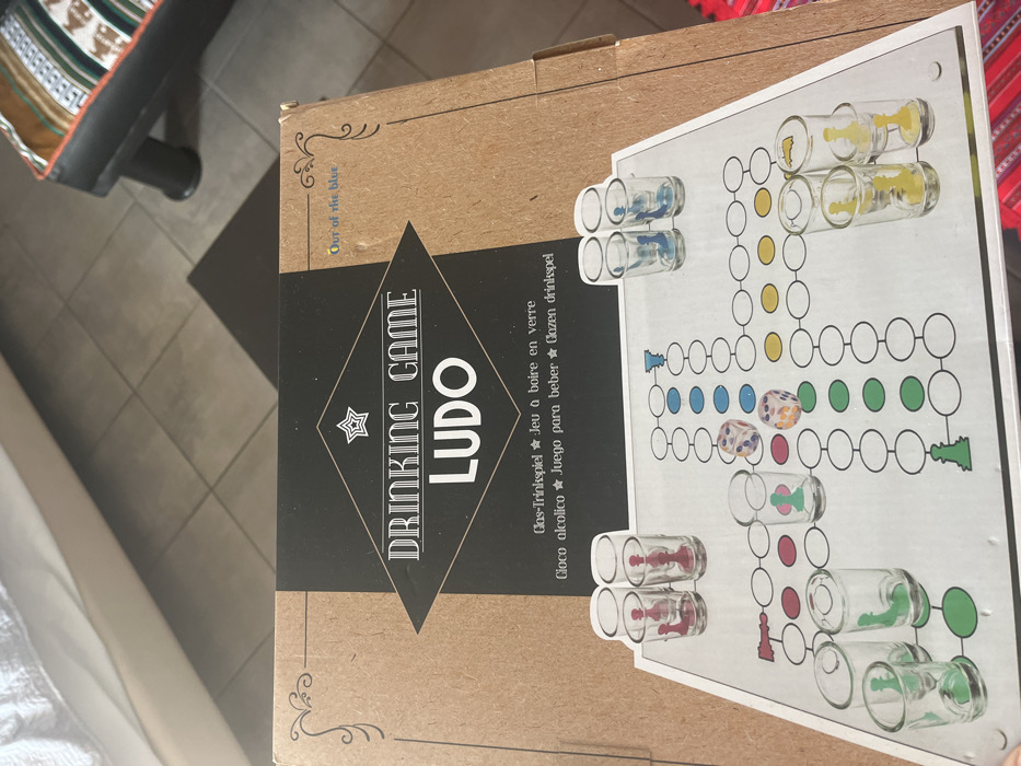 The image shows a cardboard box on a tile floor, likely indoors, with the lid slightly open. The box contains a "Drinking Game Ludo" game, designed for playing with shot glasses. The box is made of brown cardboard and has a black and white label with the game name and a depiction of a game board featuring a simplified version of Ludo, with a black and white checkered pattern, coloured circles, and five shot glasses arranged in a row on the top left side of the board, with a star in the center. The box is lying on a white bed with a white blanket. There is an orange and black patterned rug on the floor to the right. This is a common game, found in many locations, but the coordinates provided, 40.627, 24.575, locate the image in Limenaria, a small town on the island of Thasos, Greece. The box is the only object present in the image. There are no people in the image.