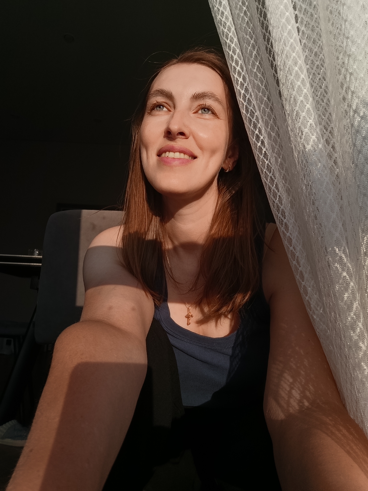 A young woman with light brown hair and blue eyes is captured in a close-up shot, bathed in the warm glow of sunset. She is looking upwards and to her left, with a gentle smile on her face, her eyes reflecting the light. She is wearing a dark blue tank top and a delicate gold cross pendant necklace. Sunlight streams in from the right side of the frame, illuminating her face and right arm, while the left side of the scene is cast in shadow. A white lace curtain hangs on the right, casting intricate patterns of light and shadow on her arm and clothing. The background appears to be an indoor setting, possibly a living room, with a dark wall and the corner of a chair visible on the left. The overall impression is one of serene contemplation and a peaceful moment captured by the natural light. The specific location within Chaikovsky, Russia, is not discernable from the visual information provided in the image.