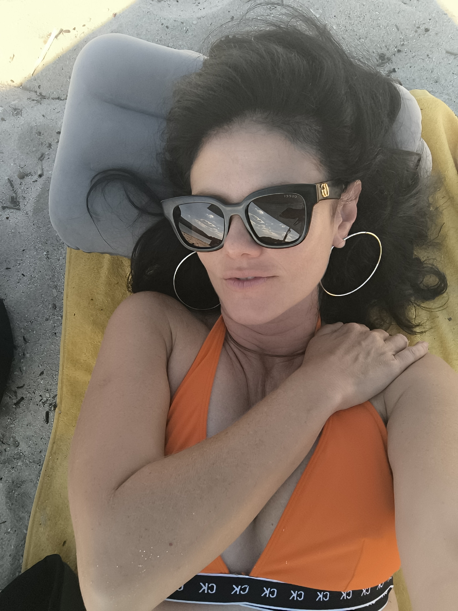 A woman lies on her back on a yellow towel and gray inflatable pillow on a sandy beach. She is wearing an orange bikini top with a black band that has "CK" repeatedly printed on it. She has dark hair, is wearing large black sunglasses with gold accents, and large silver hoop earrings. Her right arm is resting on her chest. The scene appears to be outdoors.