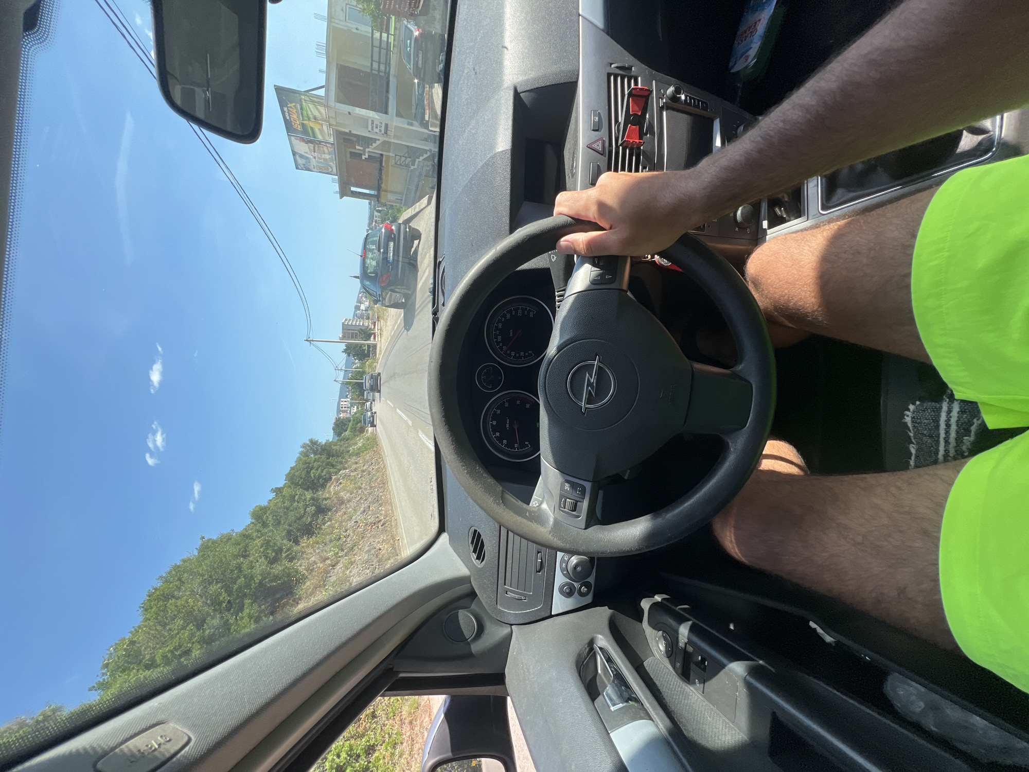 An interior shot captures a person driving an Opel vehicle along a road in Dobra Voda, Montenegro, on a sunny day. The driver's hands are on the steering wheel, and their legs are visible, wearing bright green shorts. The dashboard displays the car's instruments, and the interior of the car appears somewhat dusty. Outside the car, the road winds through a landscape with a rocky hillside covered in green vegetation on the left, and buildings and other vehicles are visible in the distance on the right. Power lines crisscross the sky.