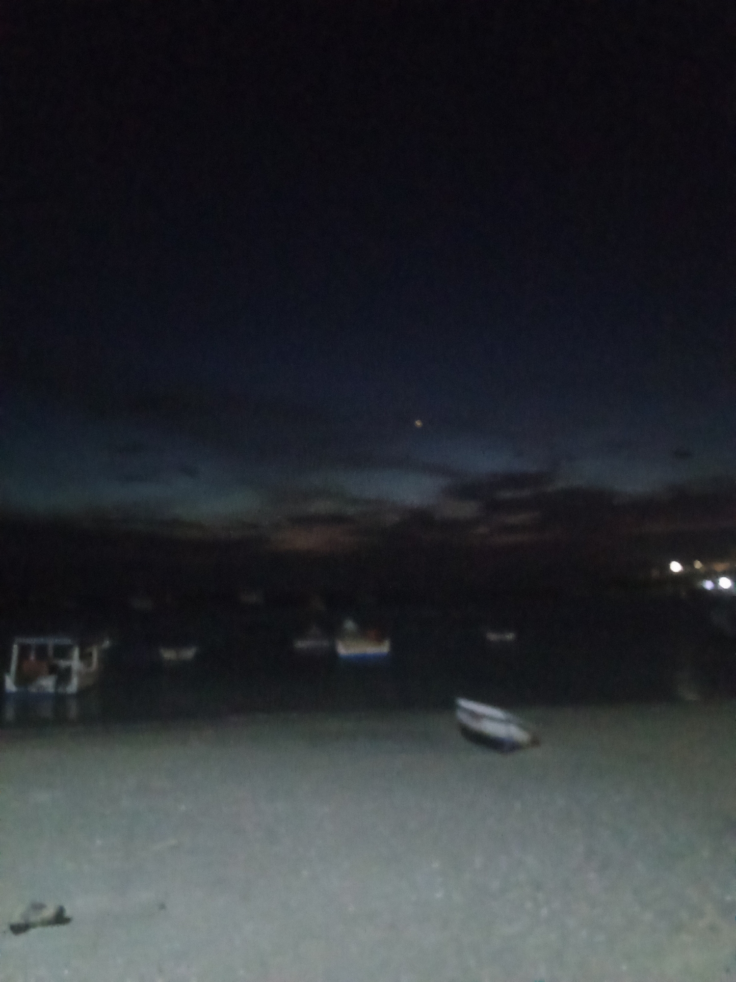 The image shows a nighttime scene at the beach in Juan Griego, Venezuela. Several boats are visible in the water, some closer to the shore. The sky is dark, with faint signs of sunset or artificial lights on the horizon. The foreground is sandy. There's a dark object on the sand. Lights are visible on the far right, possibly from buildings.
