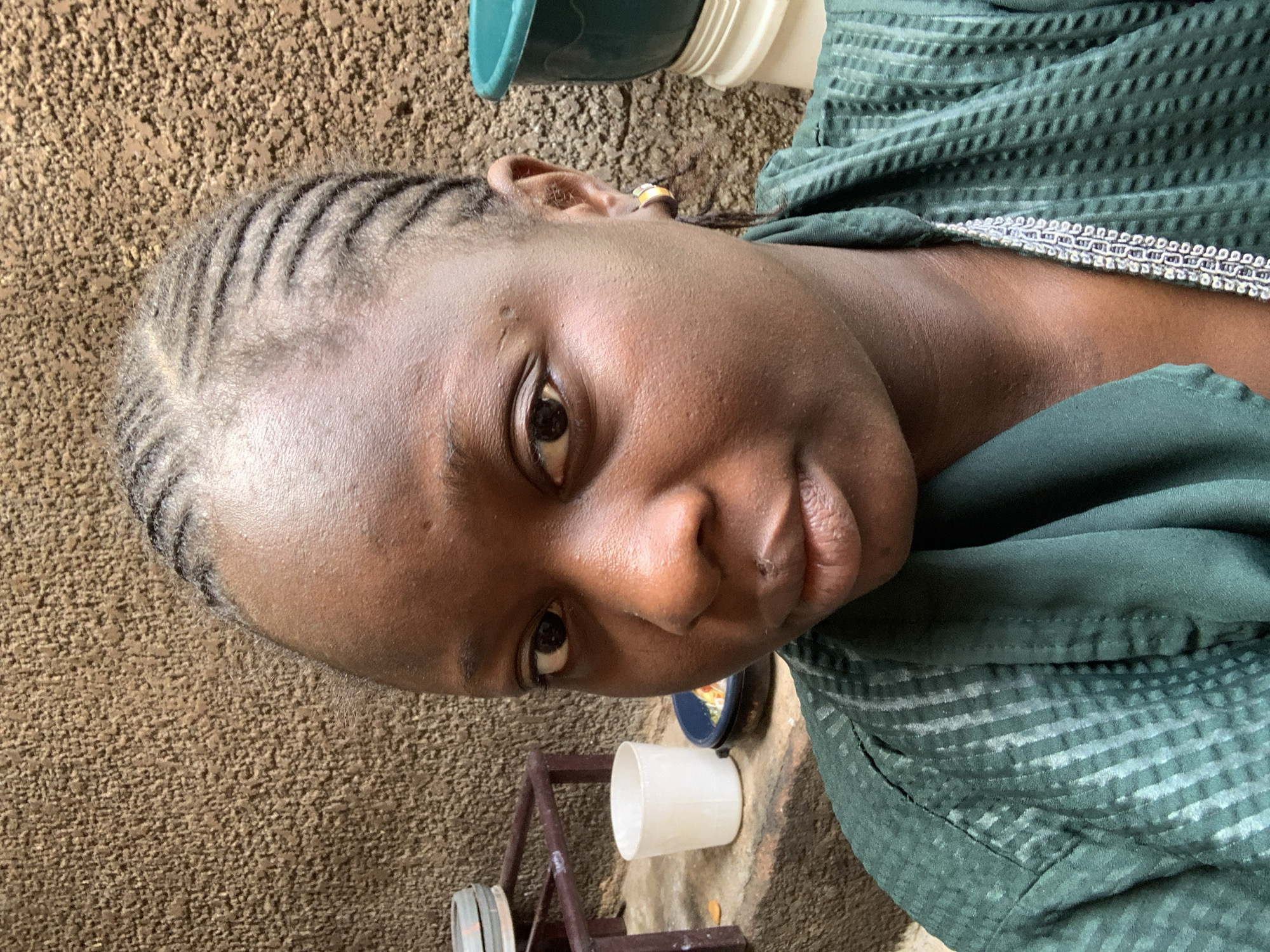 A close-up, front-facing selfie of a young Black woman with braided hair and a green robe is presented. She is positioned in front of a textured brown wall. To her left, on a ledge, are a white cup and a black bowl containing food. A green tub and a white bucket are visible to her right. Her left earlobe sports a small hoop earring with colorful embellishments. The lighting is natural and originates from the front, casting soft shadows.