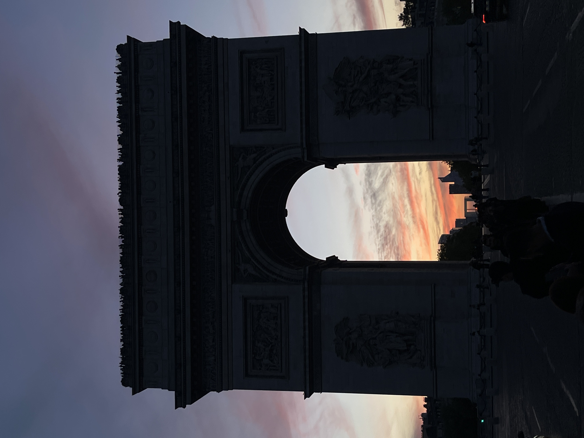 The image shows the Arc de Triomphe in Paris, France at dusk. The sky behind the arch is pink and orange from the sunset. The arch itself is a dark silhouette. People are present along the street. There are trees and buildings beyond the arch. A vehicle is visible on the right.