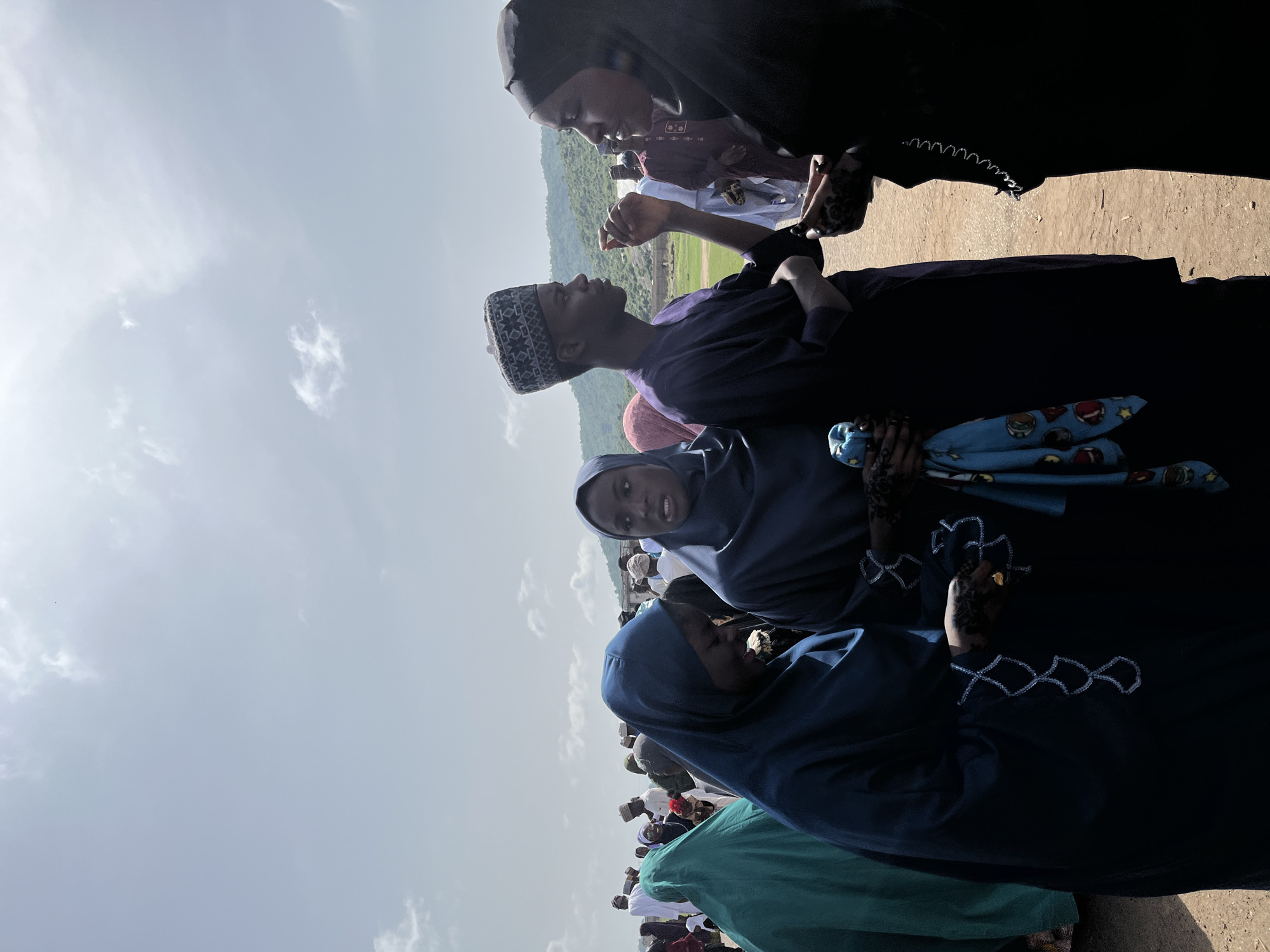 The image captures a group of individuals, likely gathered outdoors in the city of Naraguta Hausawa, Nigeria. The setting appears to be a public space with a view of a hillside in the distance. Several people are wearing traditional Muslim attire, including hijabs and other head coverings. The scene suggests a community gathering, though the specific event is unclear. Some individuals are interacting with each other, while others are standing and looking in various directions. The sky is partly cloudy, and the overall lighting indicates daylight. The foreground includes a mix of people wearing different colors and styles of clothing, and some have decorative henna patterns on their hands.