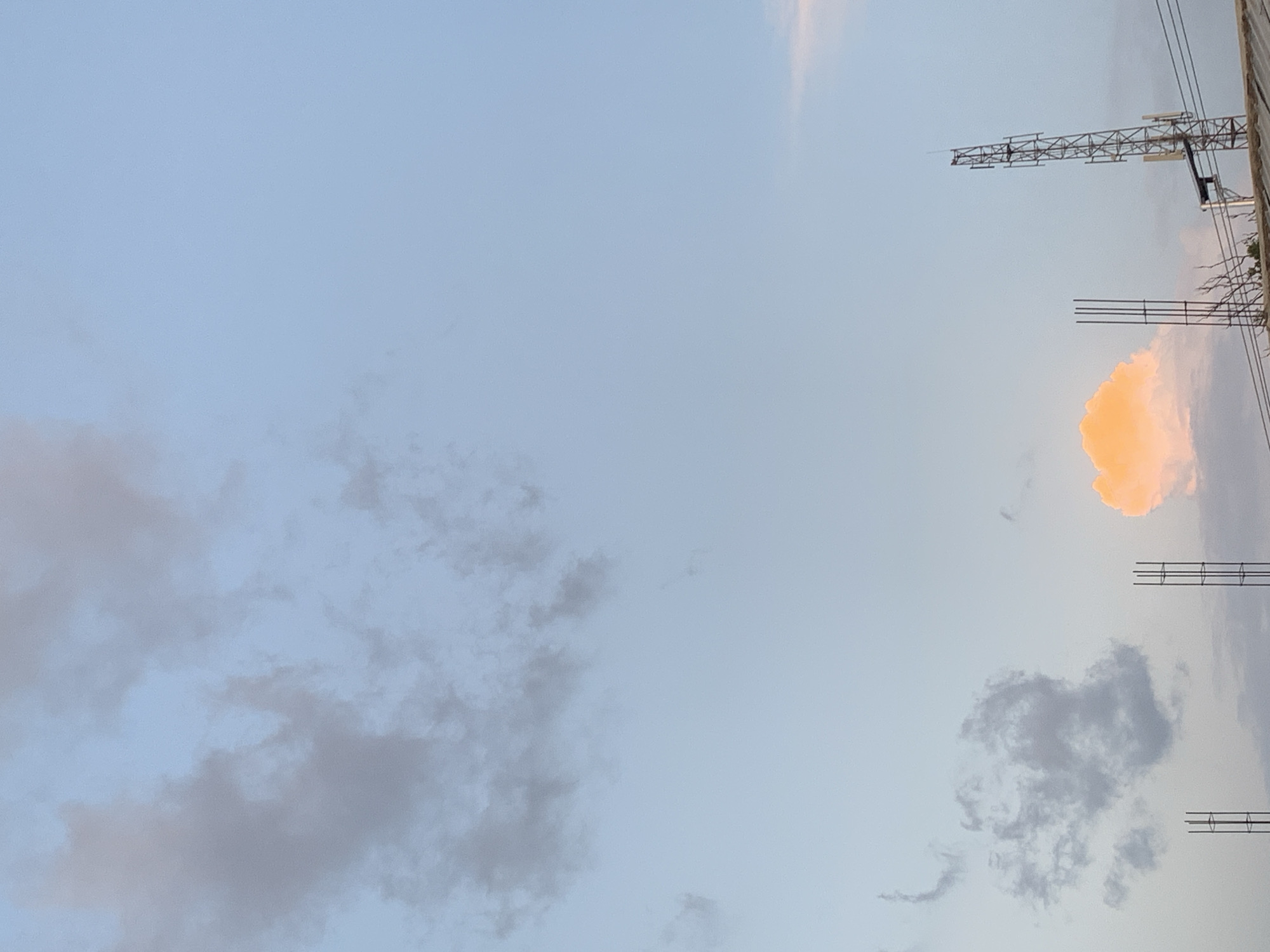 The image shows a daytime view of the sky above the New Stadium in Nigeria. The sky is predominantly light blue with scattered clouds. A cluster of grey clouds is visible on the left side of the image. On the right side, there is a partially illuminated orange cloud and above it a metallic tower structure. Metal poles are scattered along the right side of the image.