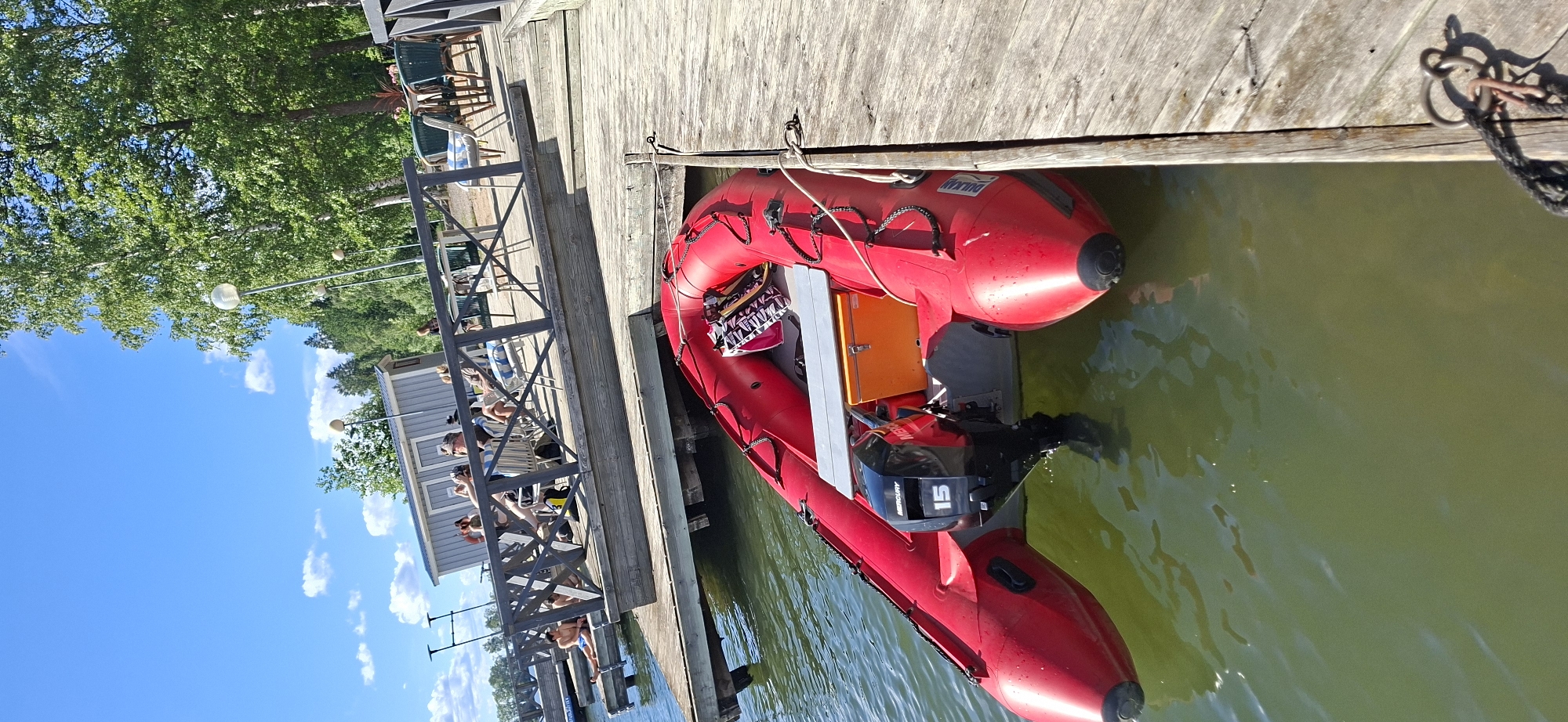 A red inflatable boat with a Mercury 15 horsepower outboard motor is tied to a wooden dock. The boat is partially submerged in murky green water. The dock is made of weathered wooden planks and is lined with a wooden railing. Behind the dock, a small gray building with white trim is visible, with several people sitting in chairs on a wooden platform in front of it. Green trees with lush foliage are in the background, and the sky is bright blue with a few scattered white clouds. The scene appears to be set during the daytime, likely on a clear and sunny day. The overall impression is a casual outdoor setting, possibly a recreational area or a lakeside spot. Visible text on the boat includes "DULKAN" and "MERCURY 15".